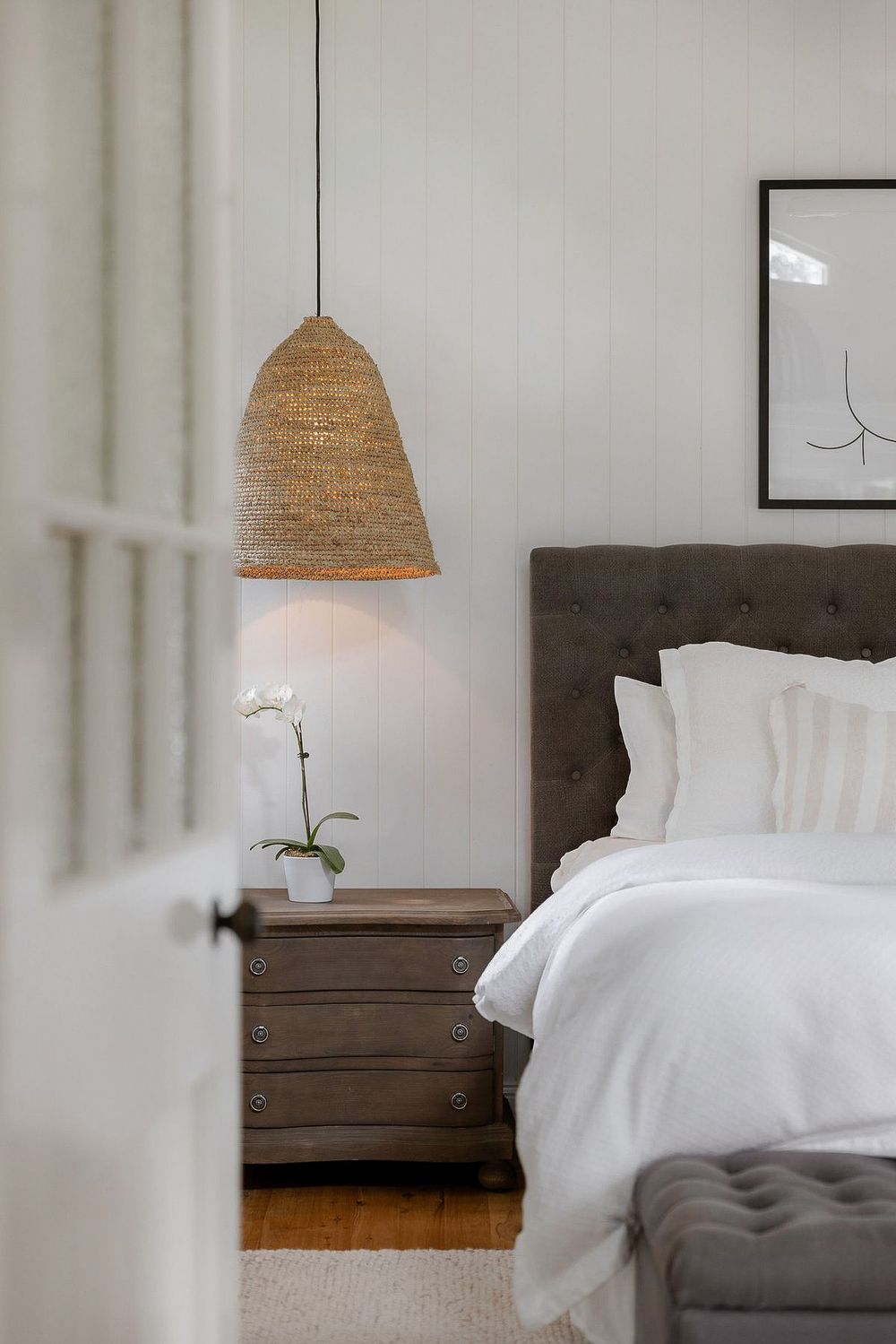 Cozy bedroom with tufted headboard, white bedding, woven pendant light, and wooden nightstand with orchid.
