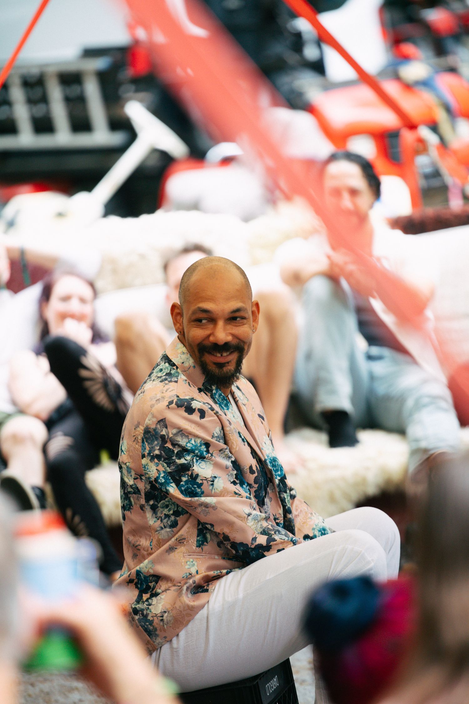 Smiling bald man with beard wearing floral jacket sits cross-legged at a colorful indoor event.