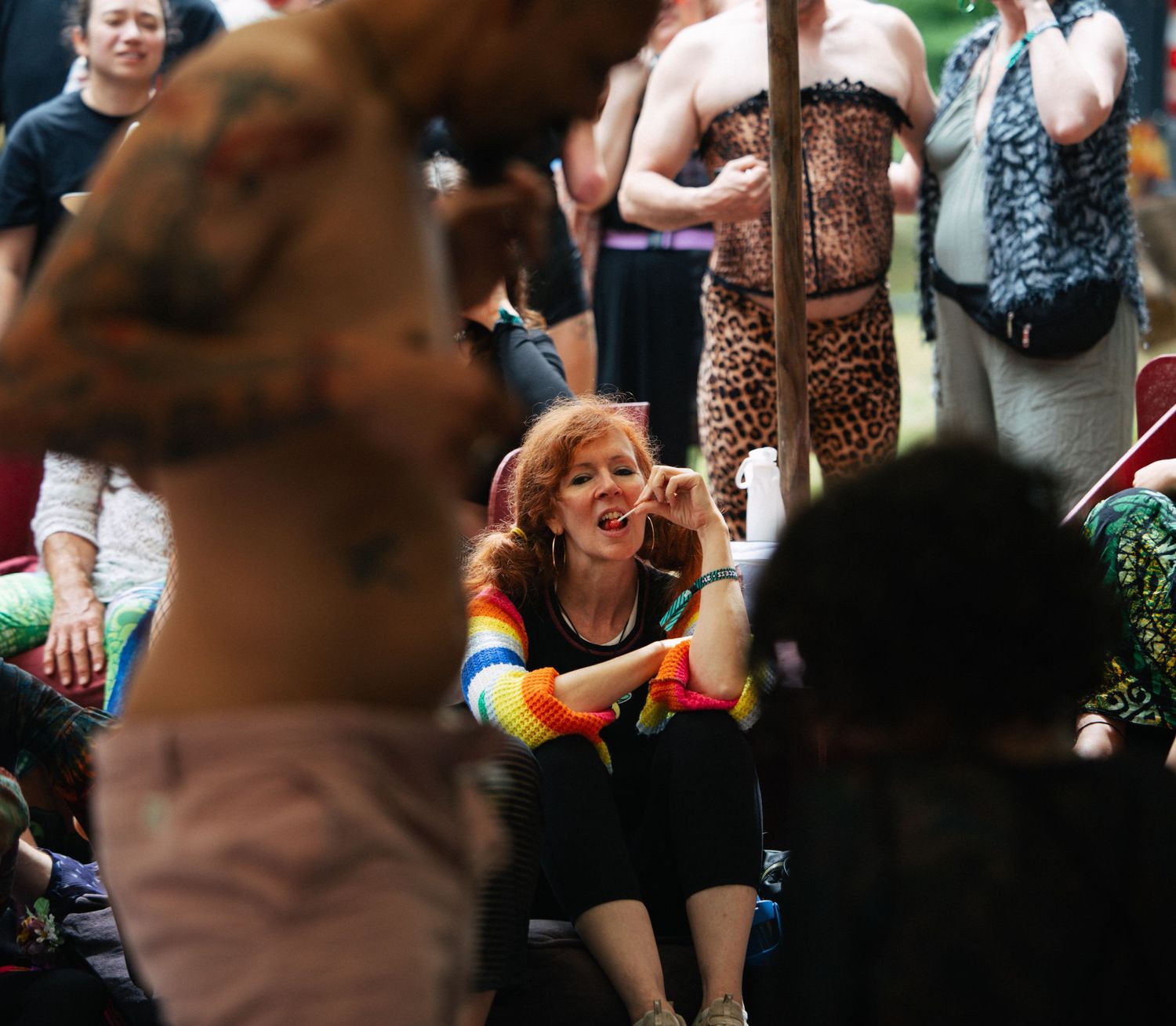 Woman in rainbow sweater sits on ground watching tattooed performer at outdoor festival event.