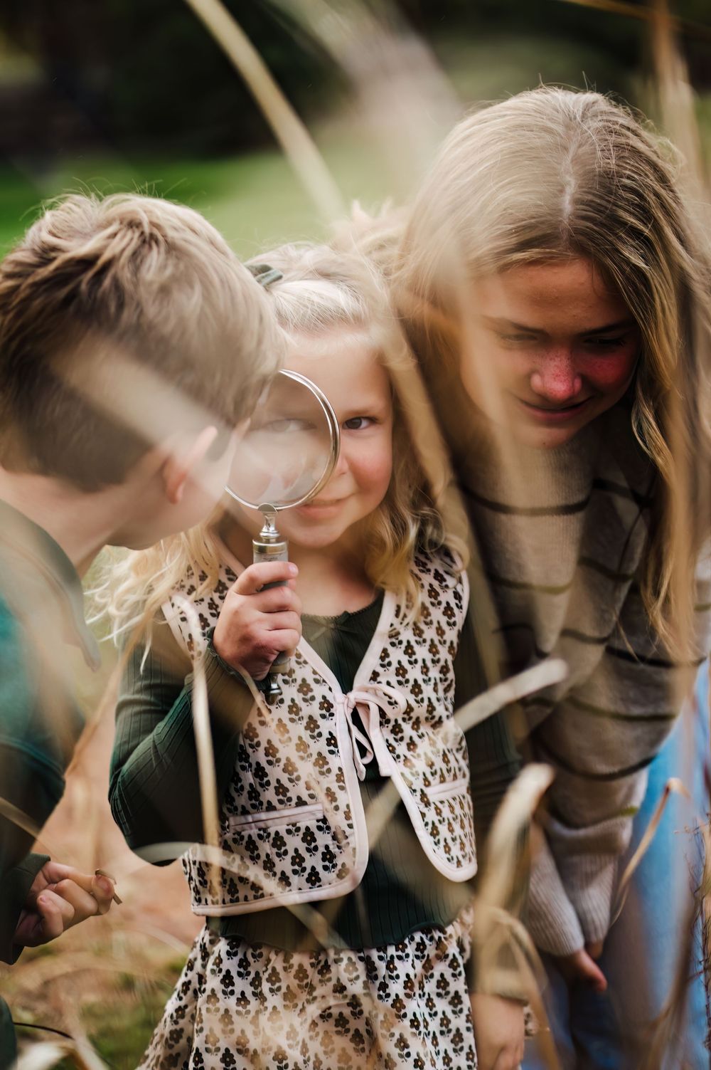 Three children exploring outdoors, with a young girl holding a magnifying glass while examining nature with her siblings.