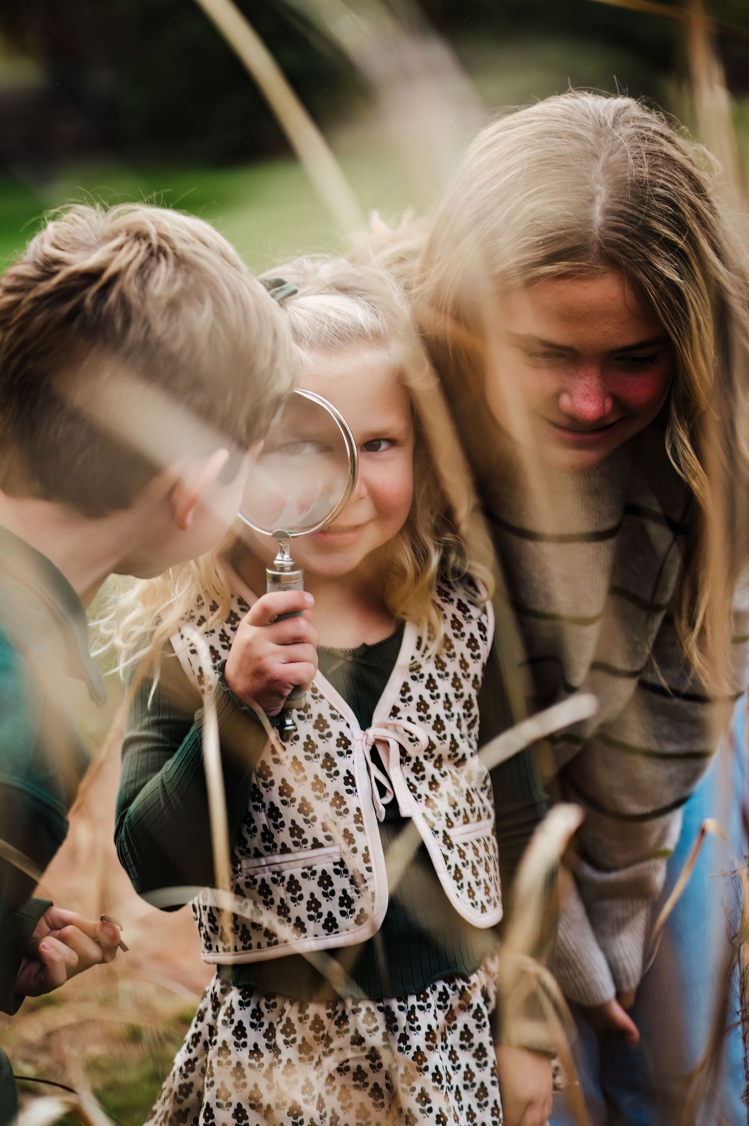 Three children exploring outdoors, with a young girl holding a magnifying glass while examining nature with her siblings.