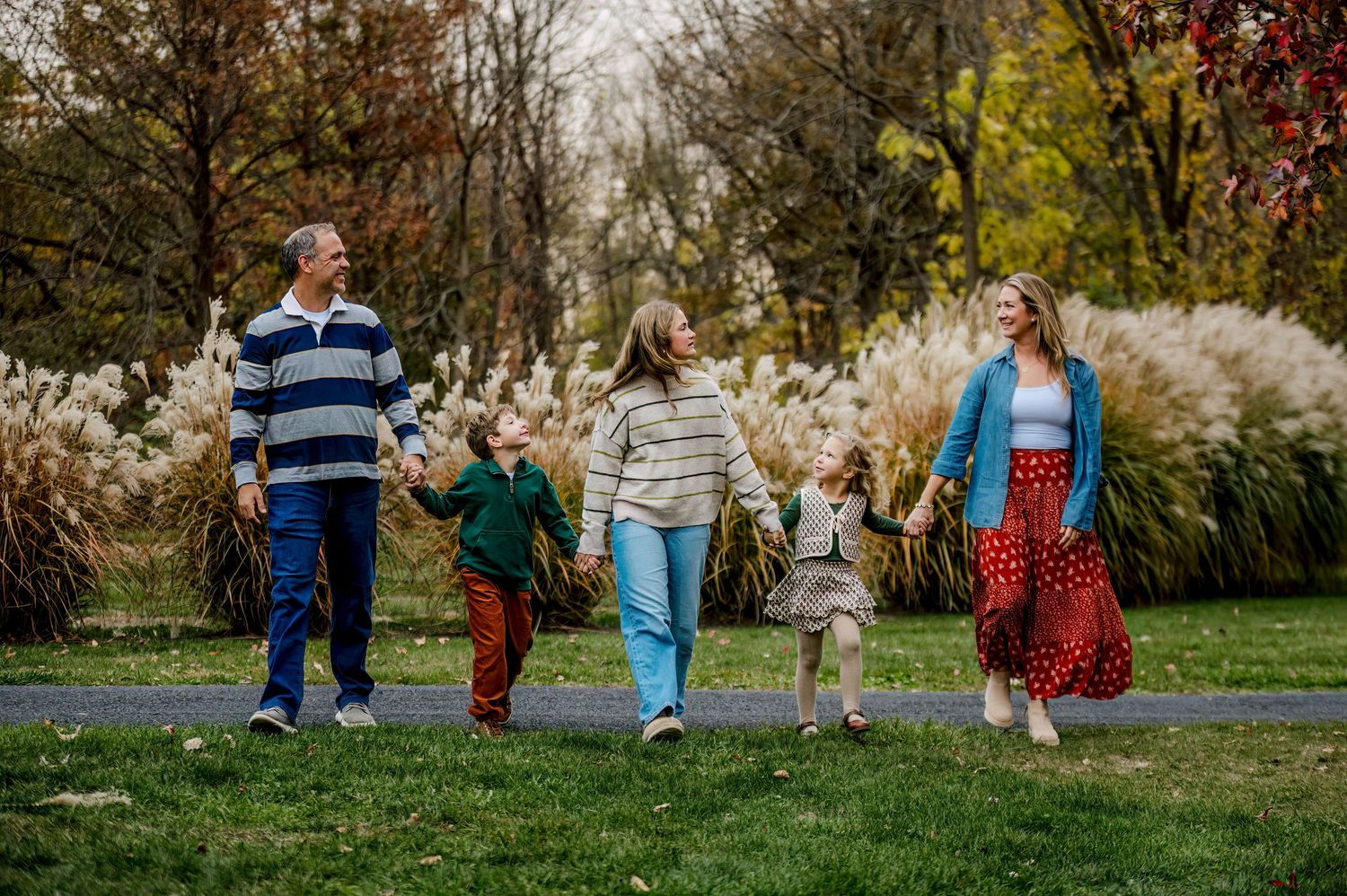 Family of four walking hand-in-hand on a park path surrounded by autumn foliage and ornamental grasses.
