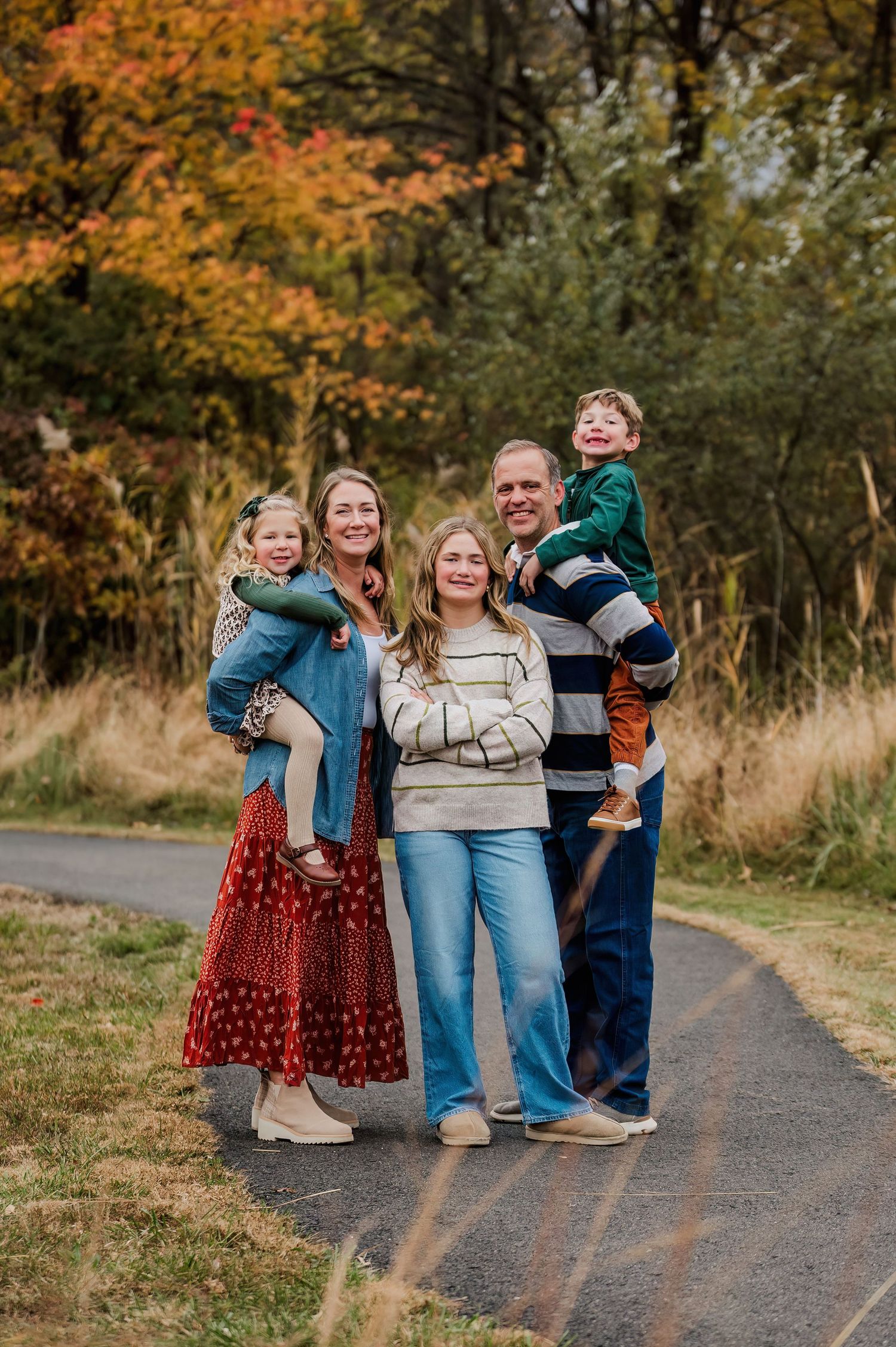 Family of five posing on a path surrounded by colorful autumn foliage, parents holding young children outdoors.