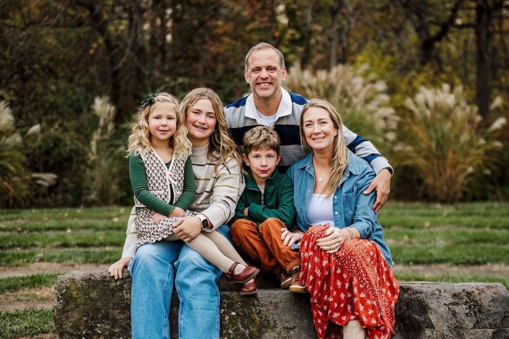 Family of five smiling together outdoors on a stone bench surrounded by lush greenery in a park setting.