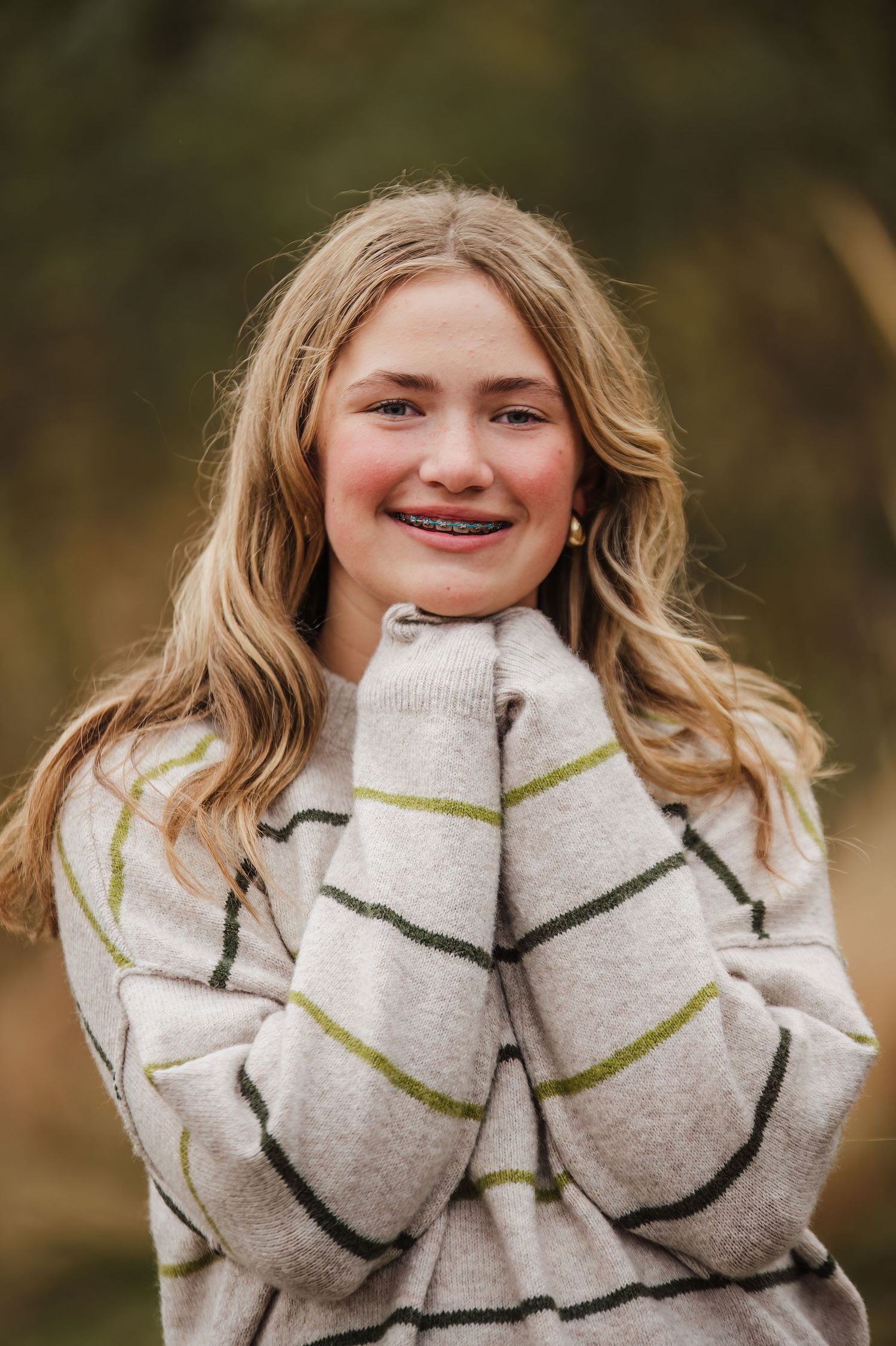 Smiling teenage girl with braces and blonde hair wearing a striped sweater, posing outdoors with hands near face.