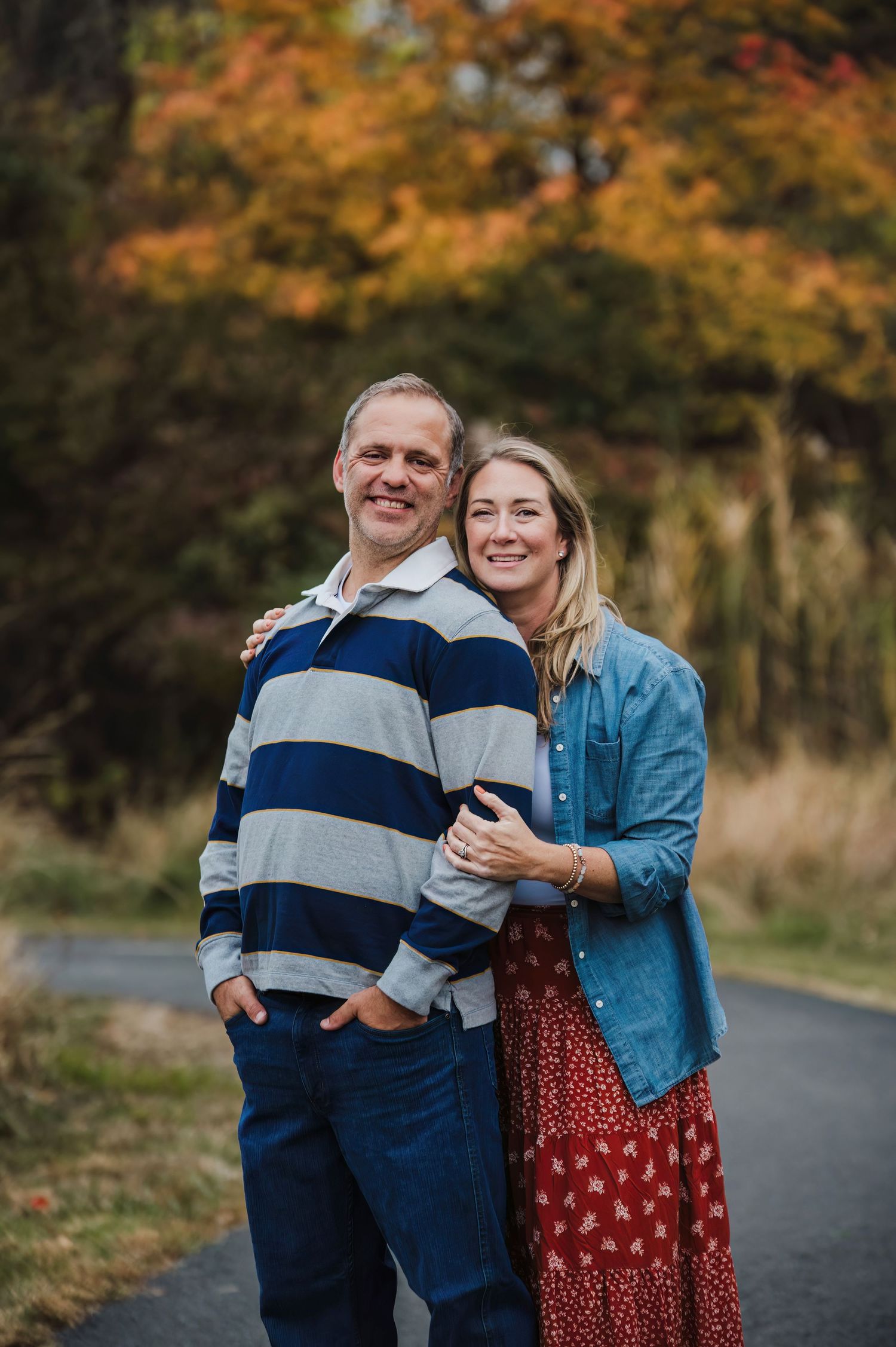 Couple smiling together on a path surrounded by colorful autumn foliage, man in striped shirt and woman in blue denim jacket.