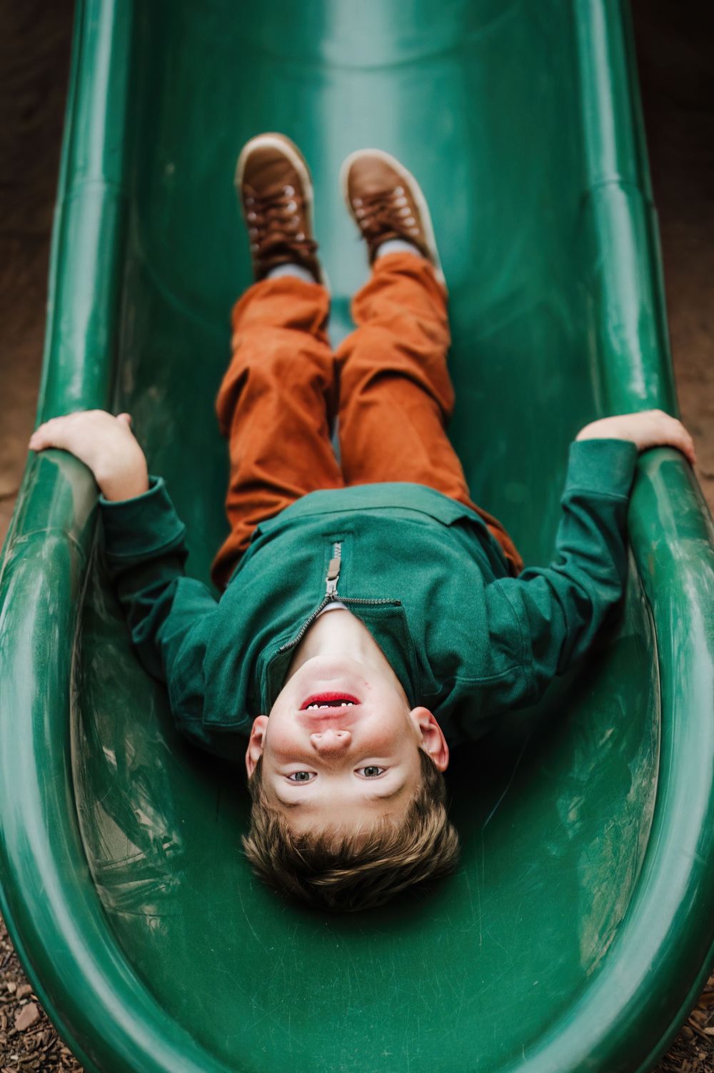Young boy in green sweatshirt and orange pants laughing while sliding down a green playground slide.