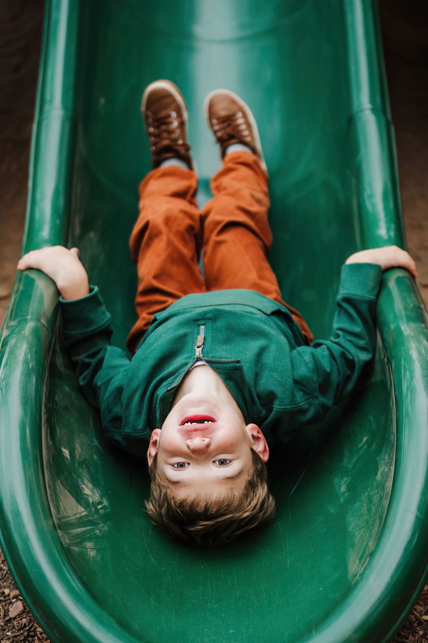 Young boy in green sweatshirt and orange pants laughing while sliding down a green playground slide.
