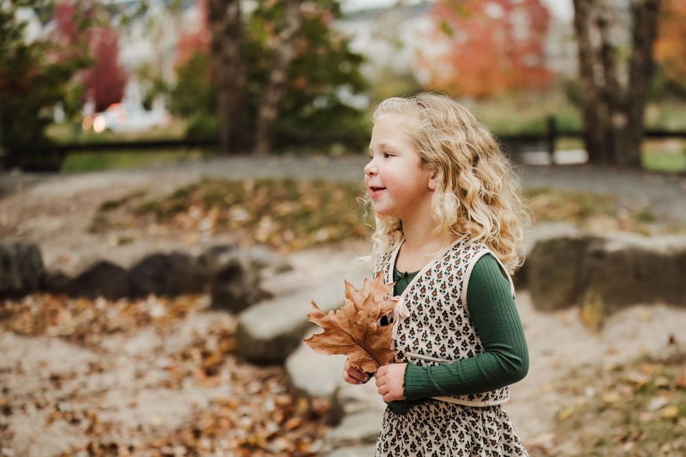 A young blonde girl holds autumn leaves in a park, wearing a patterned dress with green sleeves on a fall day.