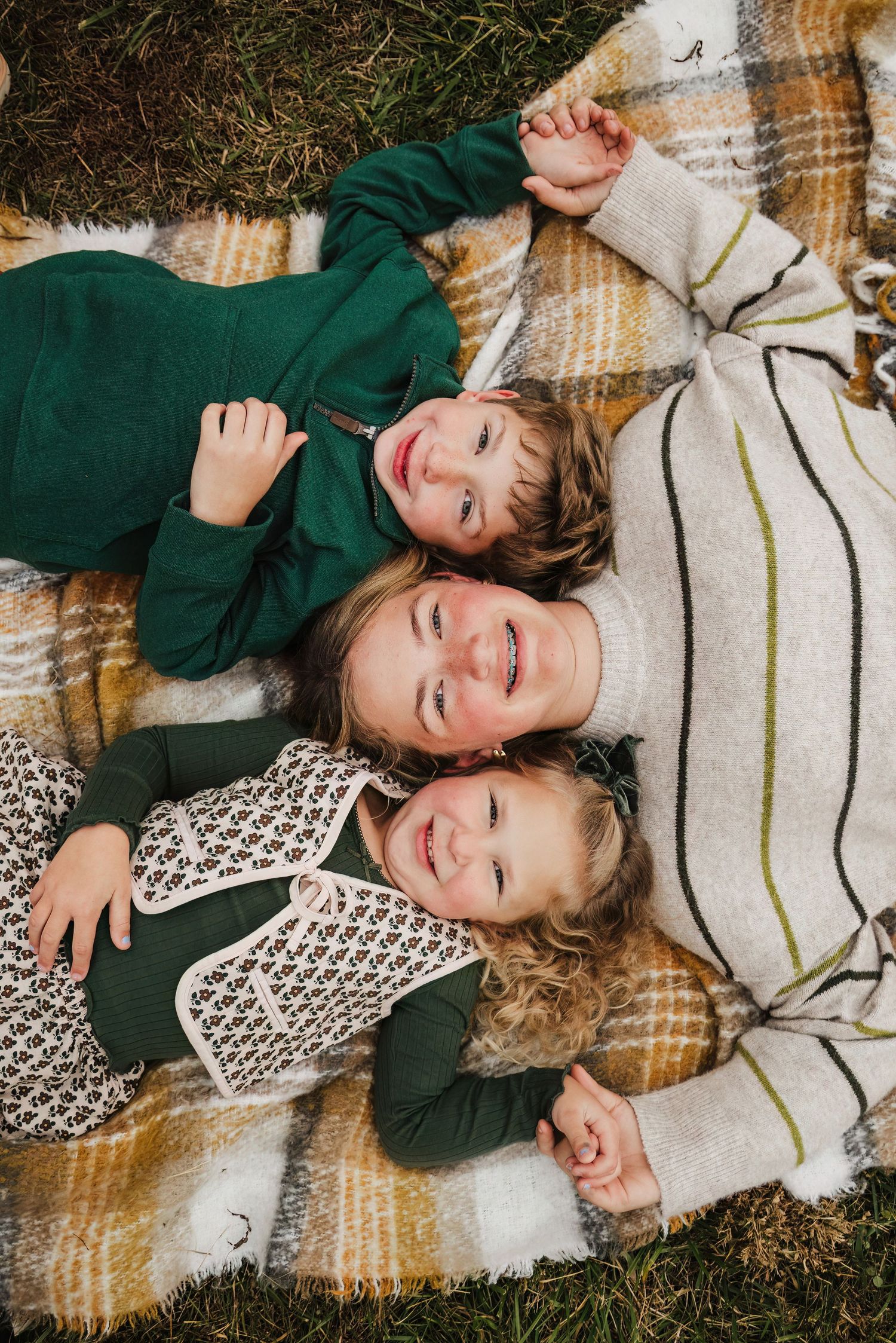 Three children lying on a plaid blanket outdoors, smiling up at the camera in green and neutral fall outfits.