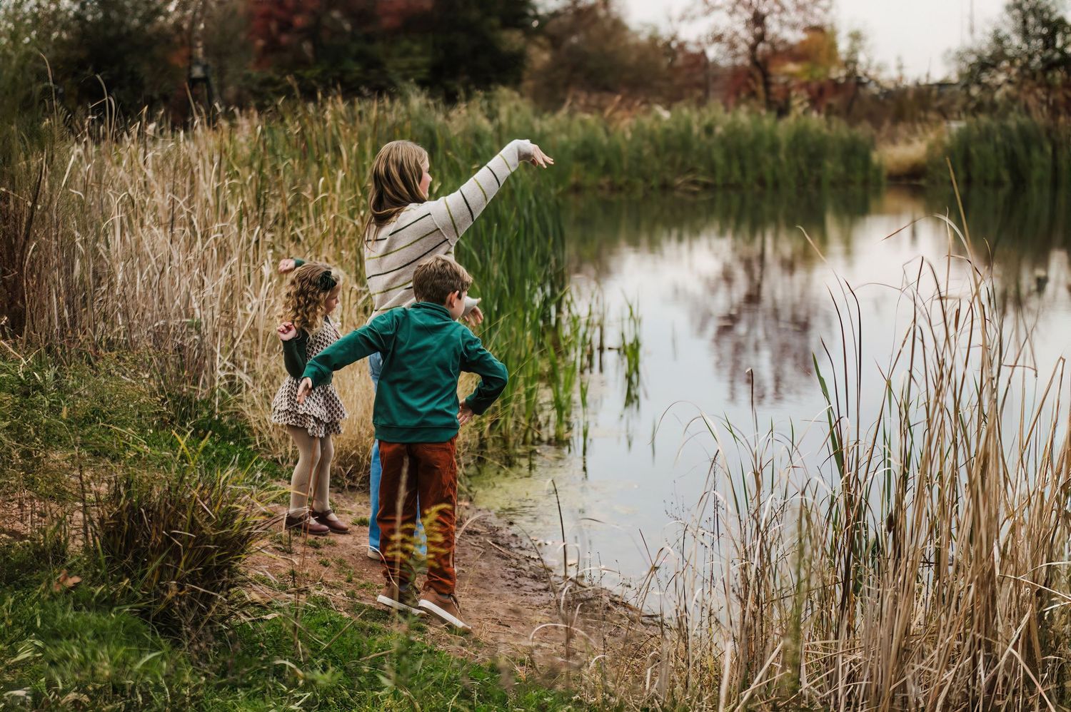 Three children stand at the edge of a serene pond surrounded by tall reeds, with one child pointing across the water.