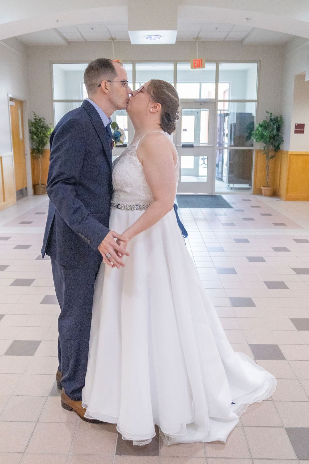 Bride and groom sharing a kiss in wedding attire inside a bright hall, white gown and navy suit.