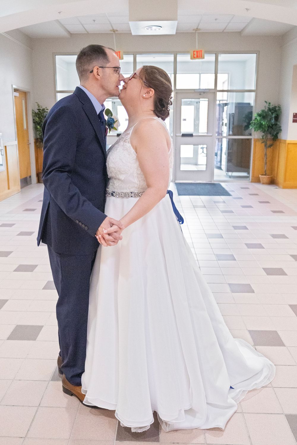 A bride and groom share a kiss while holding hands in a bright indoor venue, wearing wedding attire.
