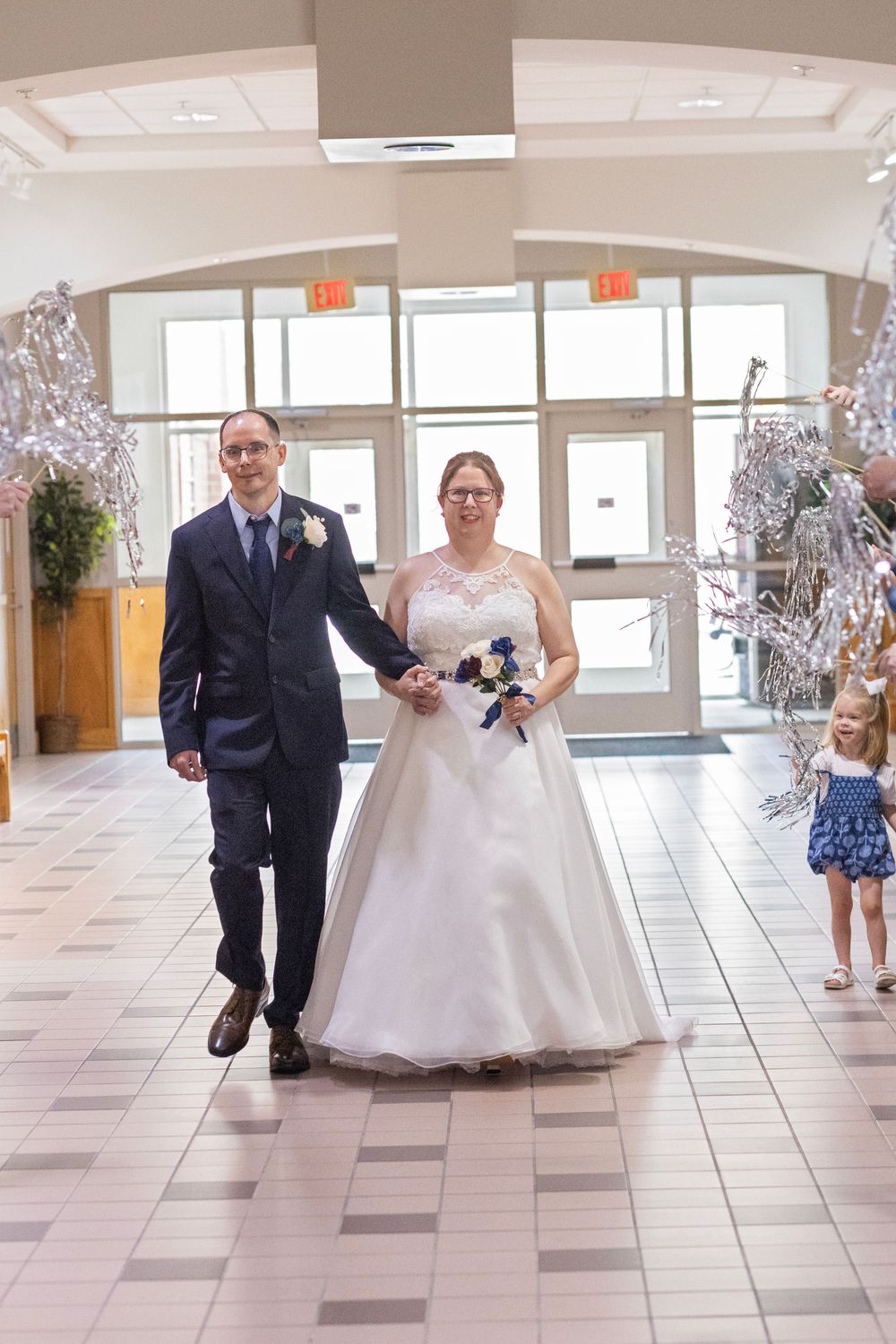 Bride and groom walk hand-in-hand in a decorated wedding venue, bride holding a bouquet, with a young girl in blue nearby.