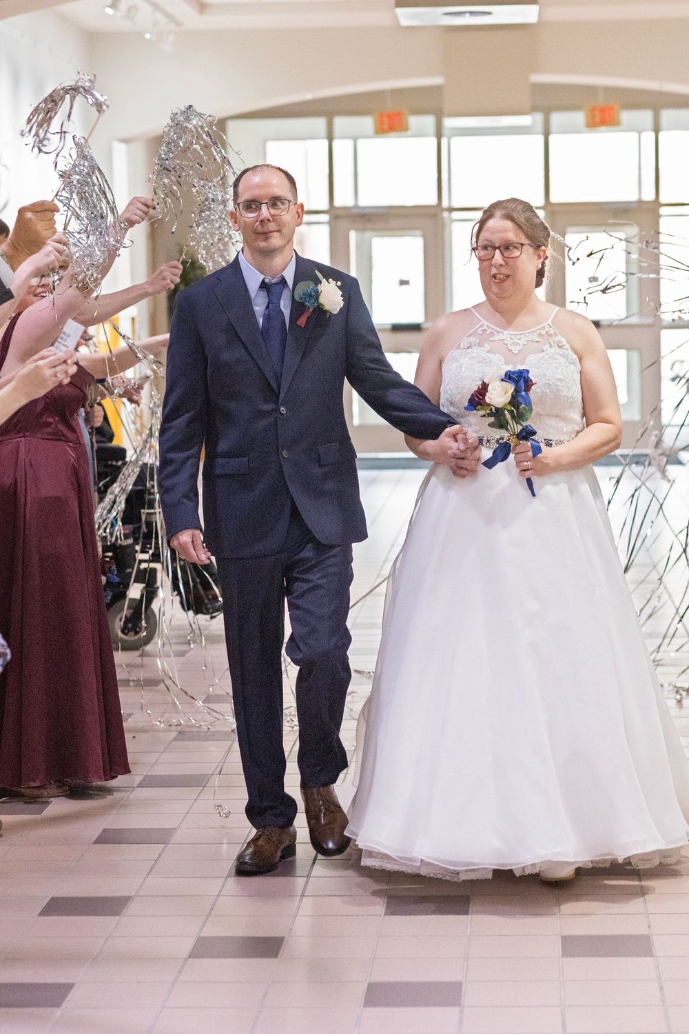 Bride and groom walk hand-in-hand through a celebratory wedding reception exit with guests waving silver streamers.