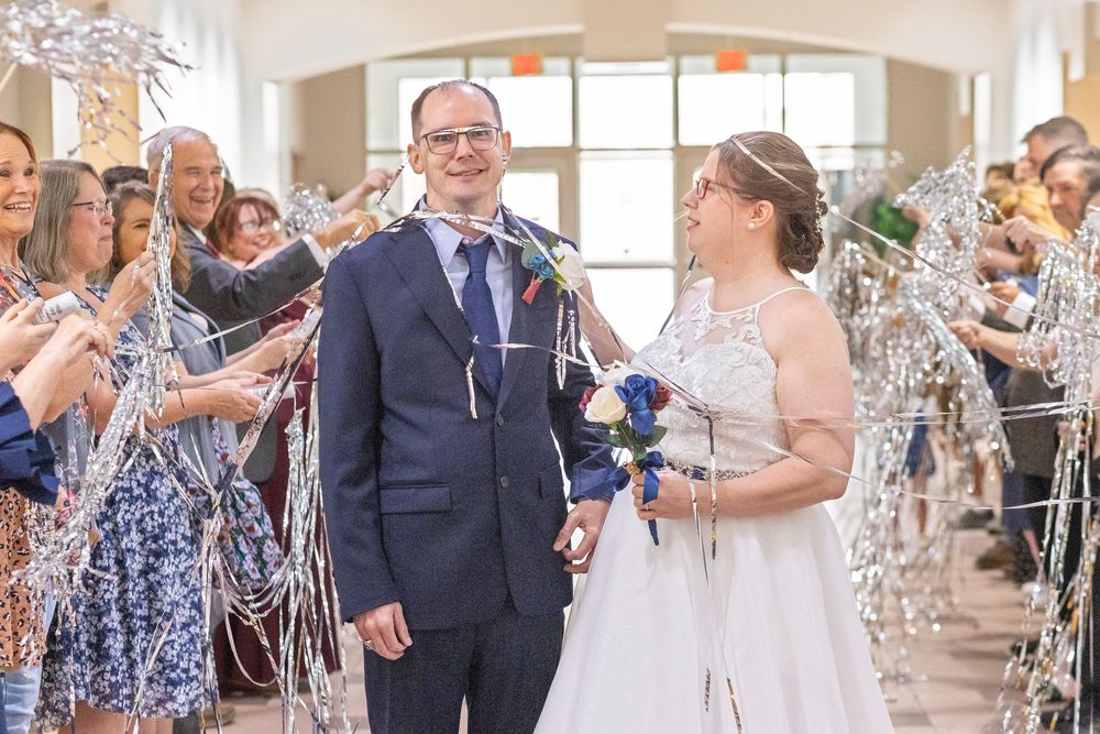 Bride and groom walk through cheering guests waving silver streamers during joyful wedding reception exit.