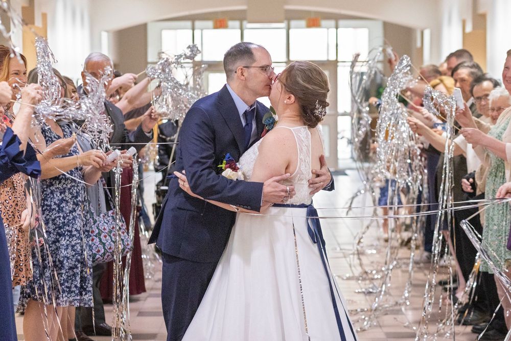 Newlyweds share a kiss during their wedding exit as guests wave ribbon wands in a bright indoor venue.