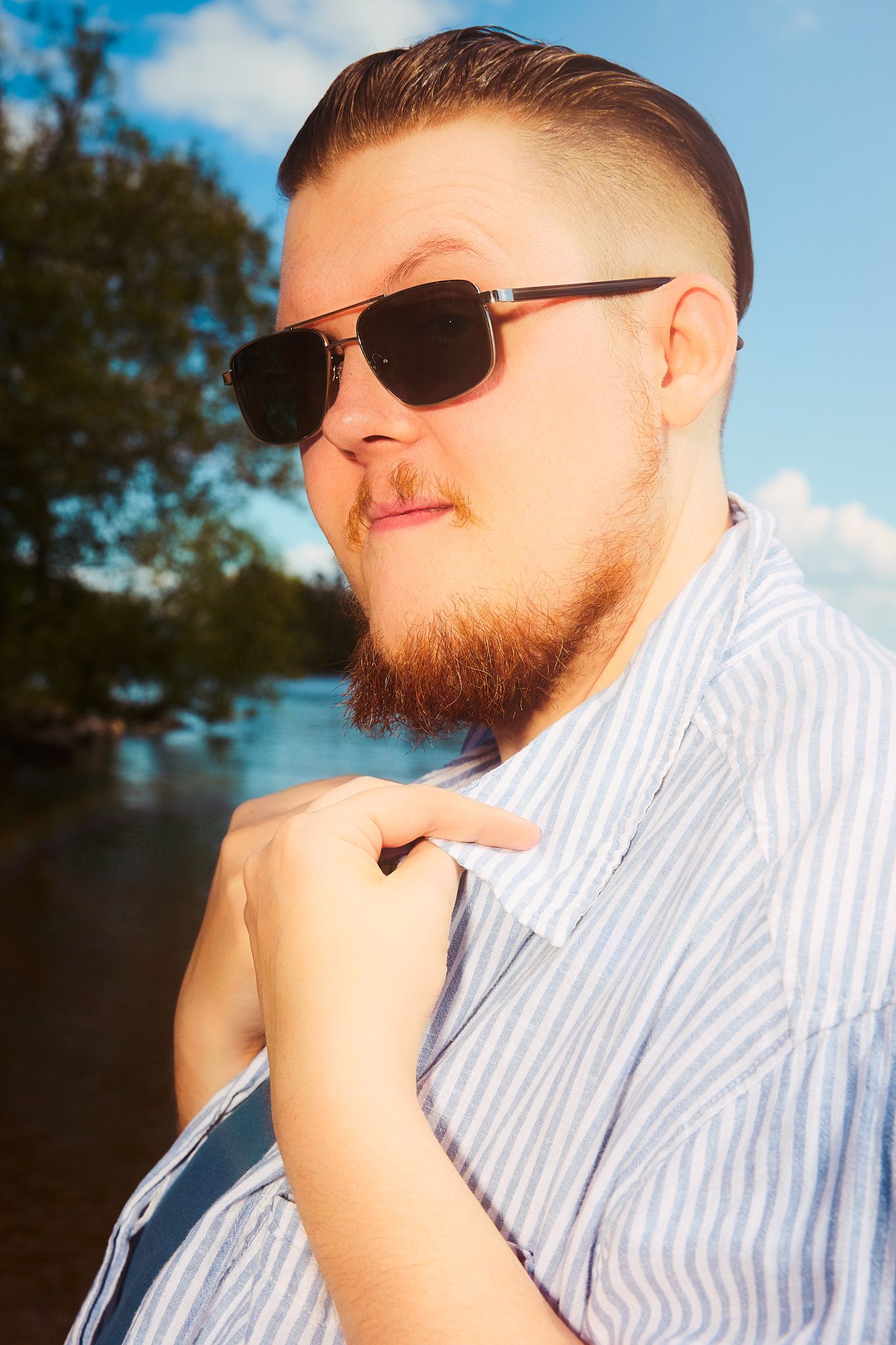 Bearded man in sunglasses and striped shirt poses outdoors near a river with trees and blue sky.