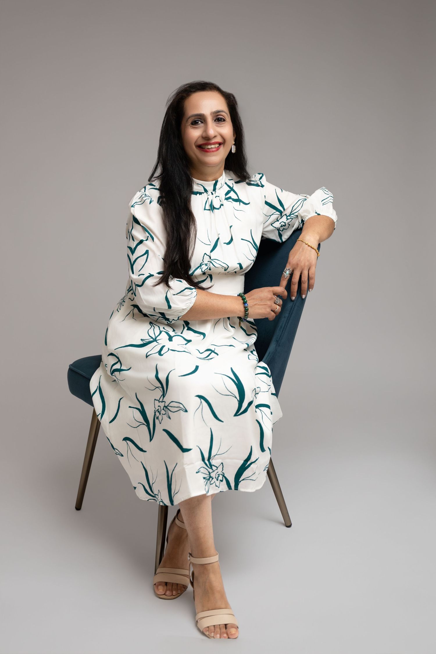 Woman in floral white and green midi dress seated on blue chair, smiling confidently in a studio setting.