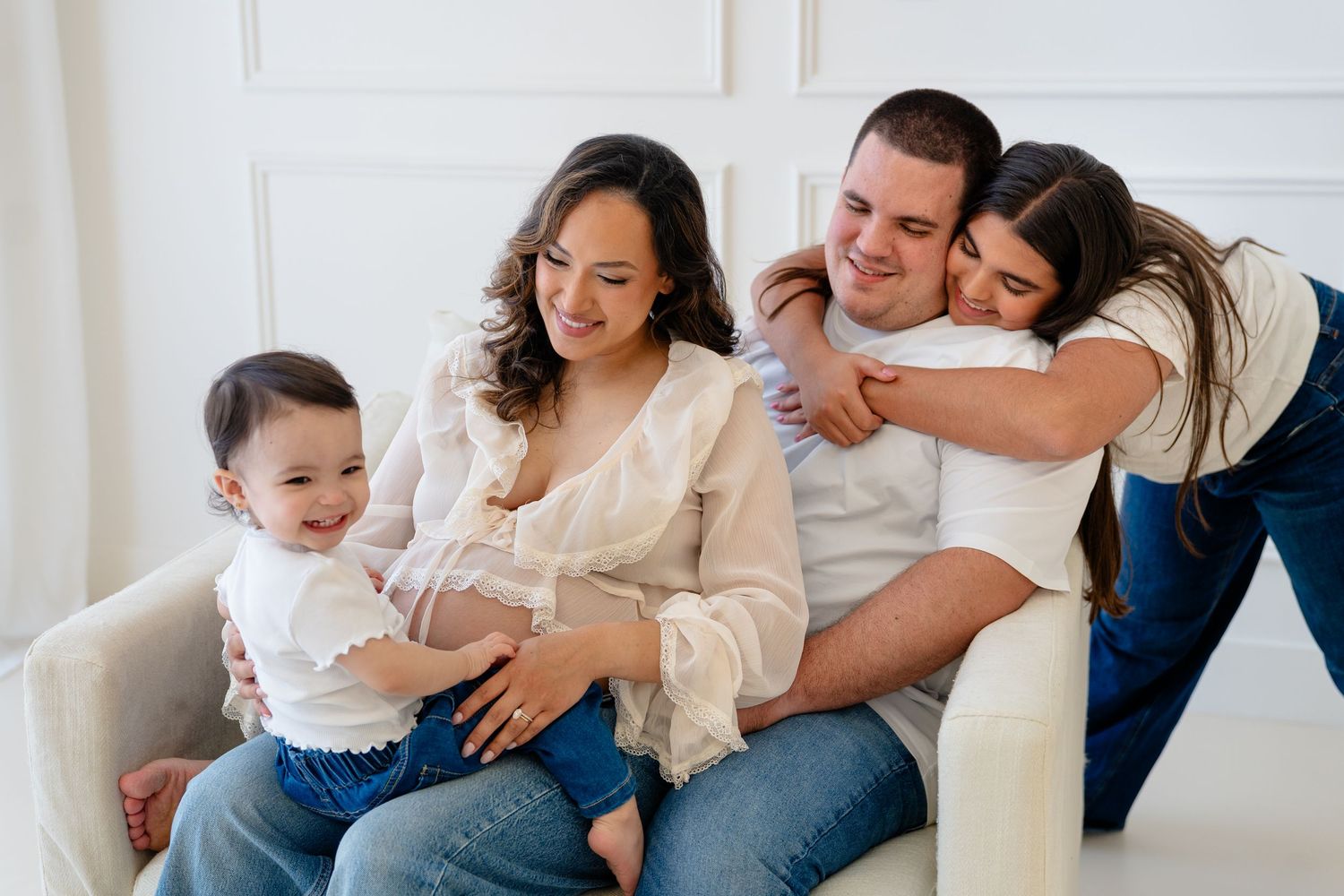 A happy family of four poses on a white couch, with a pregnant mother, father, toddler, and young daughter hugging warmly.