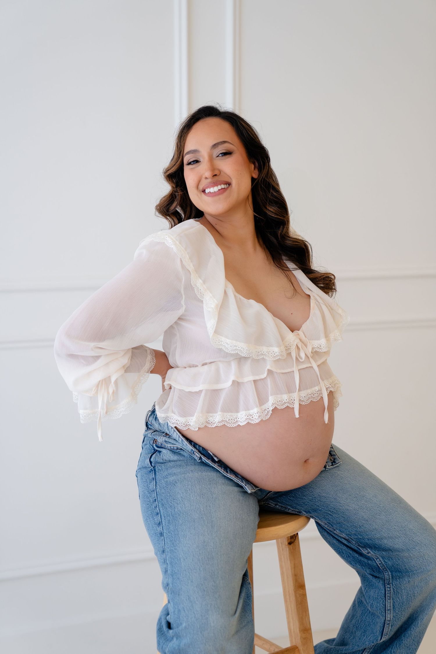 Smiling pregnant woman in white ruffled crop top and jeans sitting on stool, showing baby bump in bright studio.