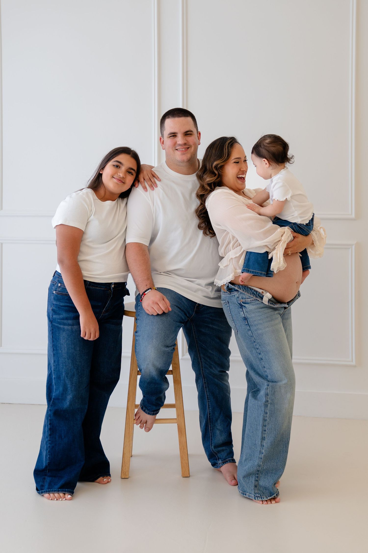 Family of four in white tops and jeans posing together in a bright studio, parents with two children smiling warmly.