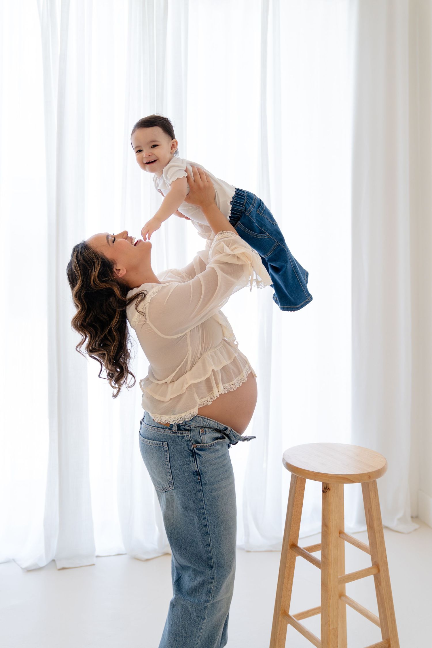 Pregnant woman in jeans lifting smiling baby in the air in a bright white studio setting.