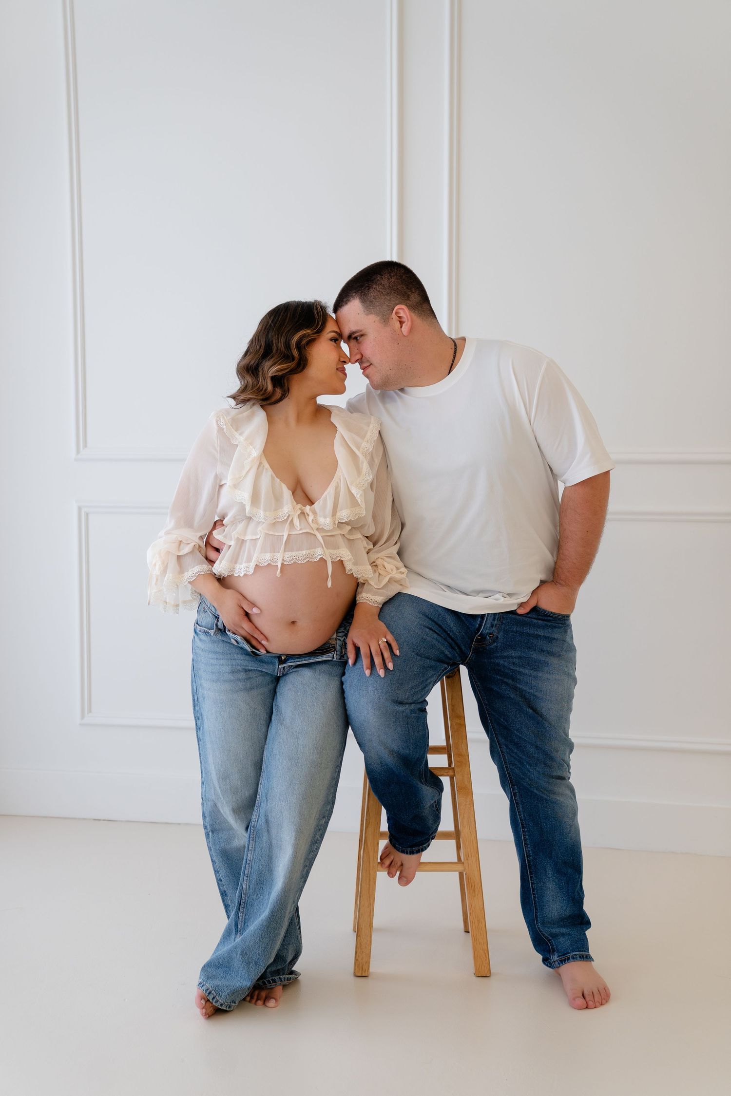 Couple sharing a tender moment, pregnant woman in sheer top and jeans with partner in white tee, posed against white wall.