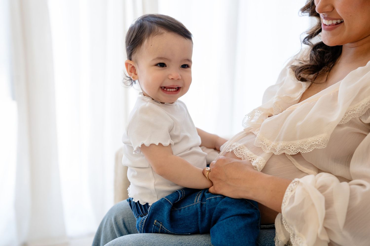 Smiling baby in white top and jeans sitting on mother's lap in bright white room.