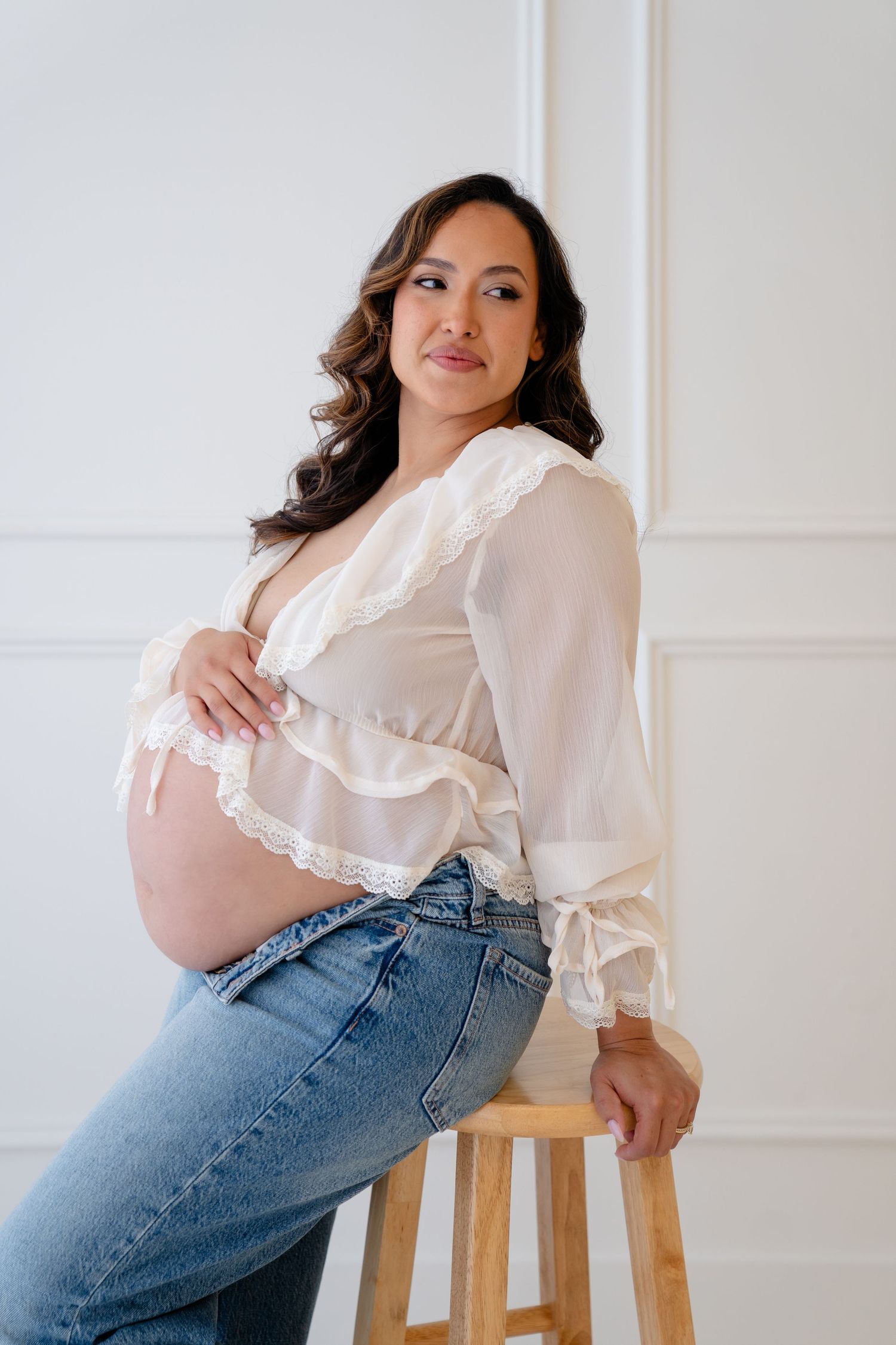 Pregnant woman in white lace top and jeans sitting on wooden stool, cradling baby bump in bright studio.