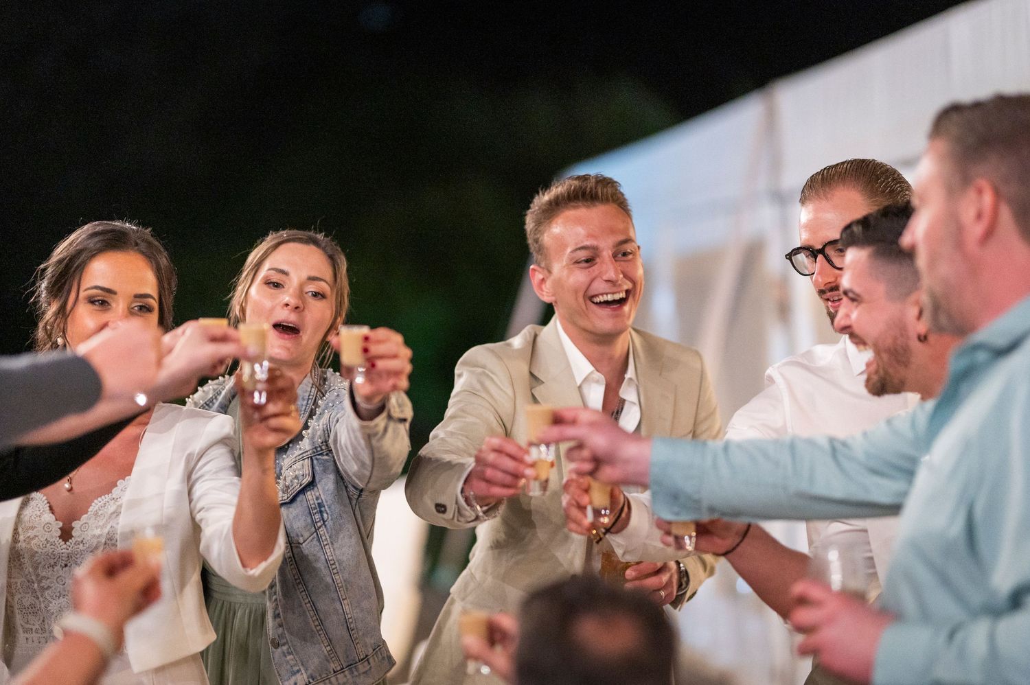 Group of friends laughing and toasting with drinks at an outdoor evening wedding celebration.