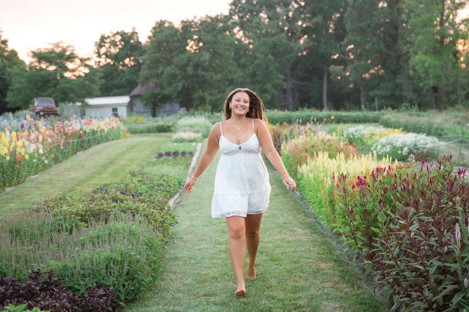 Woman in white dress walking through colorful garden rows at sunset.