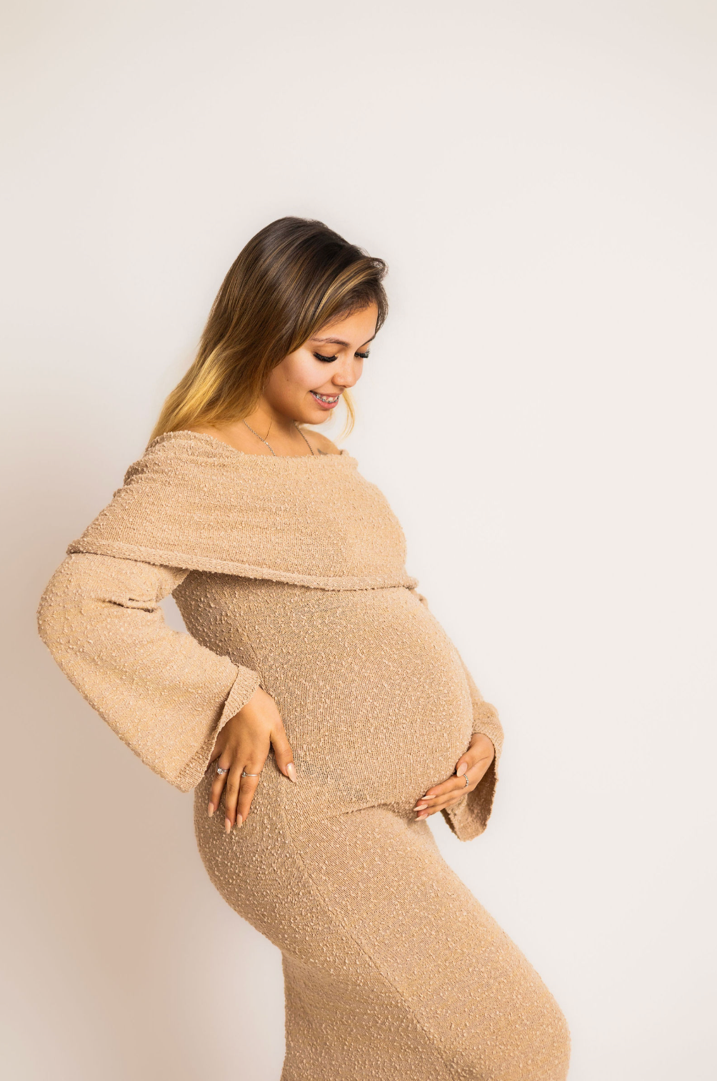 Pregnant woman in beige off-shoulder knit dress cradling baby bump, smiling softly against white background.
