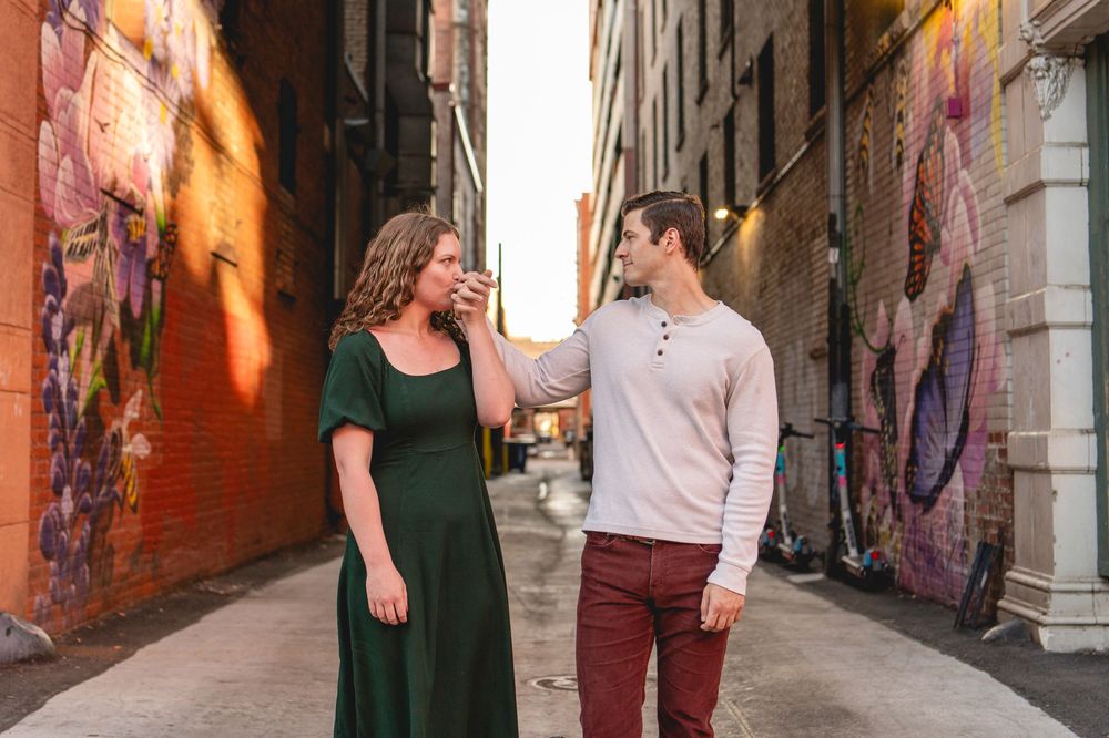 Couple posing in urban alley with colorful murals, woman in green dress, man in pink sweater and burgundy pants.