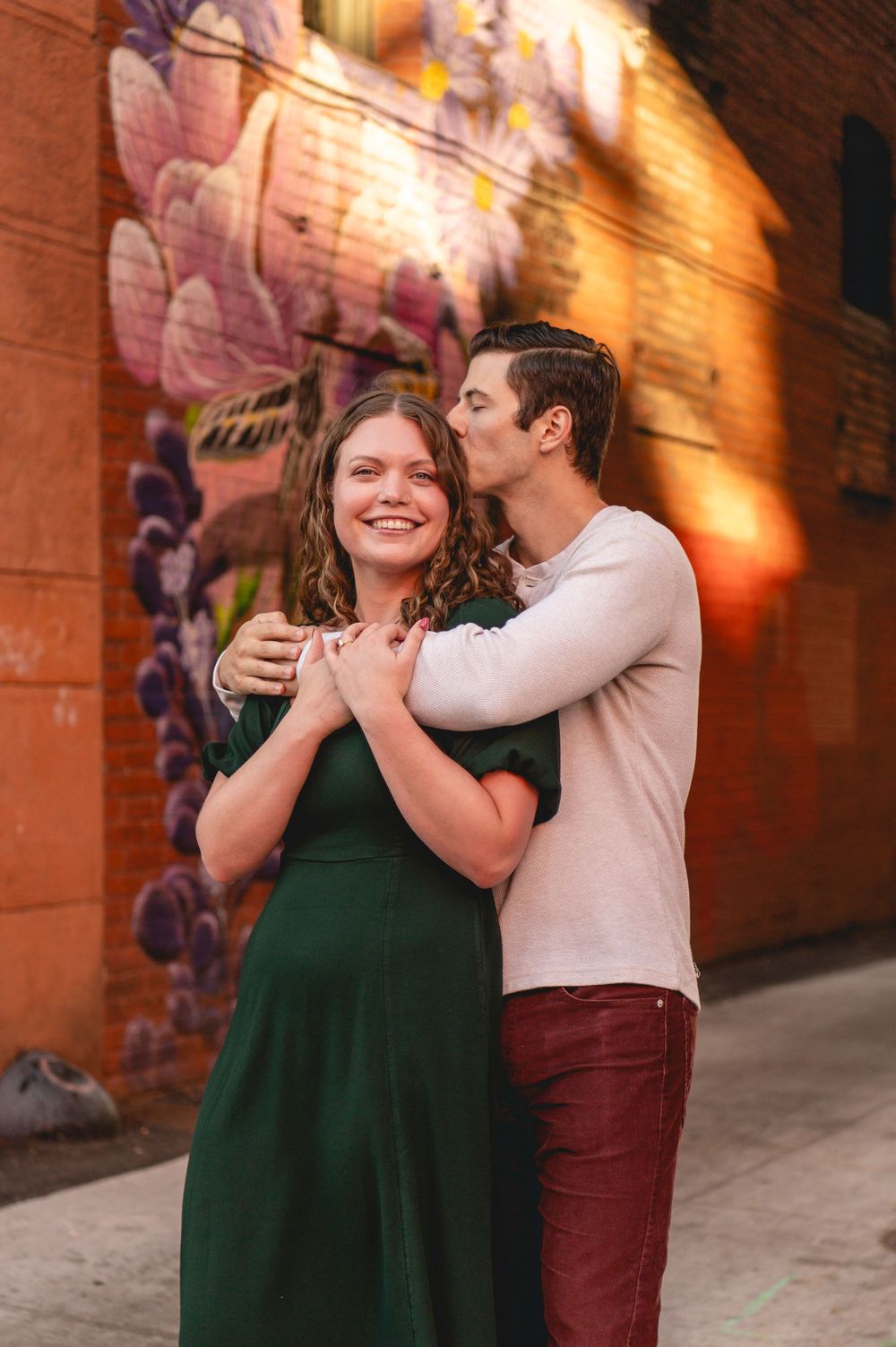 A couple embraces near a colorful floral mural, woman in green dress smiling as man kisses her cheek.