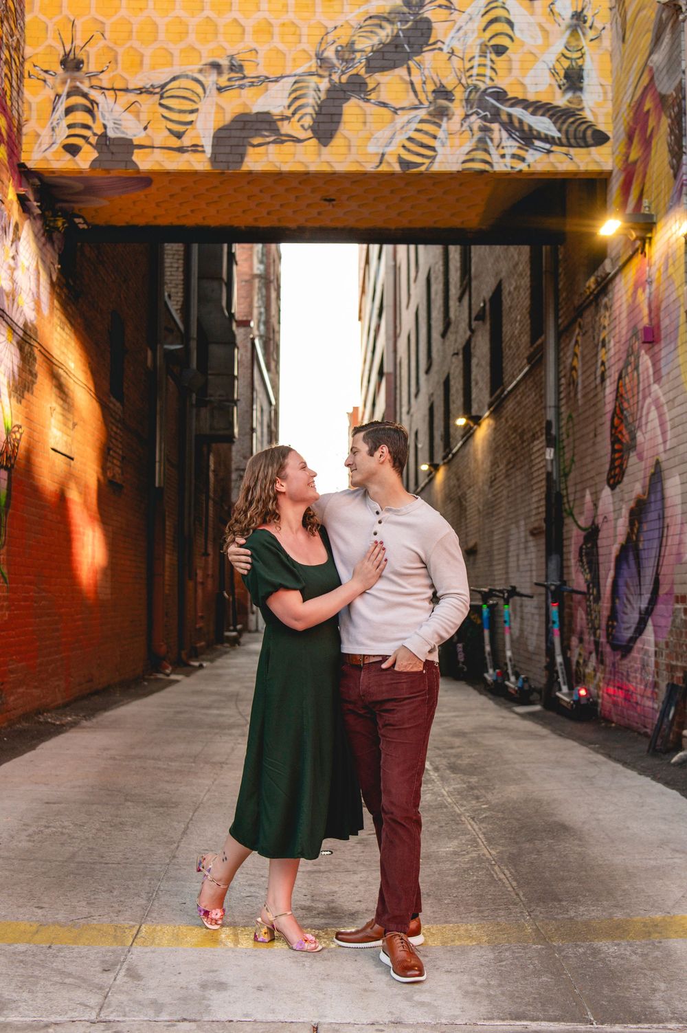 Couple embracing in a colorful urban alley with bee mural, woman in green dress, man in maroon pants.