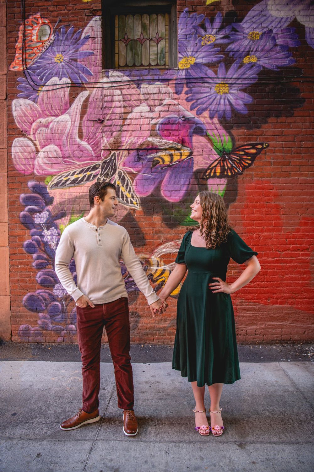 Couple holding hands smiling before a vibrant brick wall mural with flowers and butterflies in an urban setting.