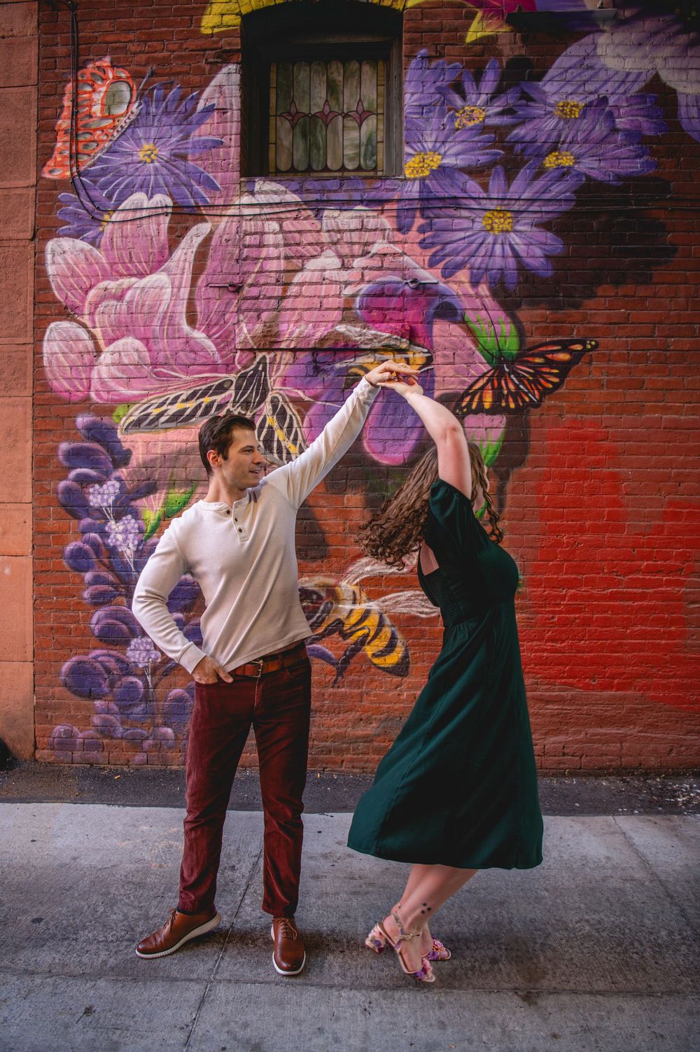 Couple dancing in front of a colorful floral mural on a brick wall, man in white shirt and woman in green dress.