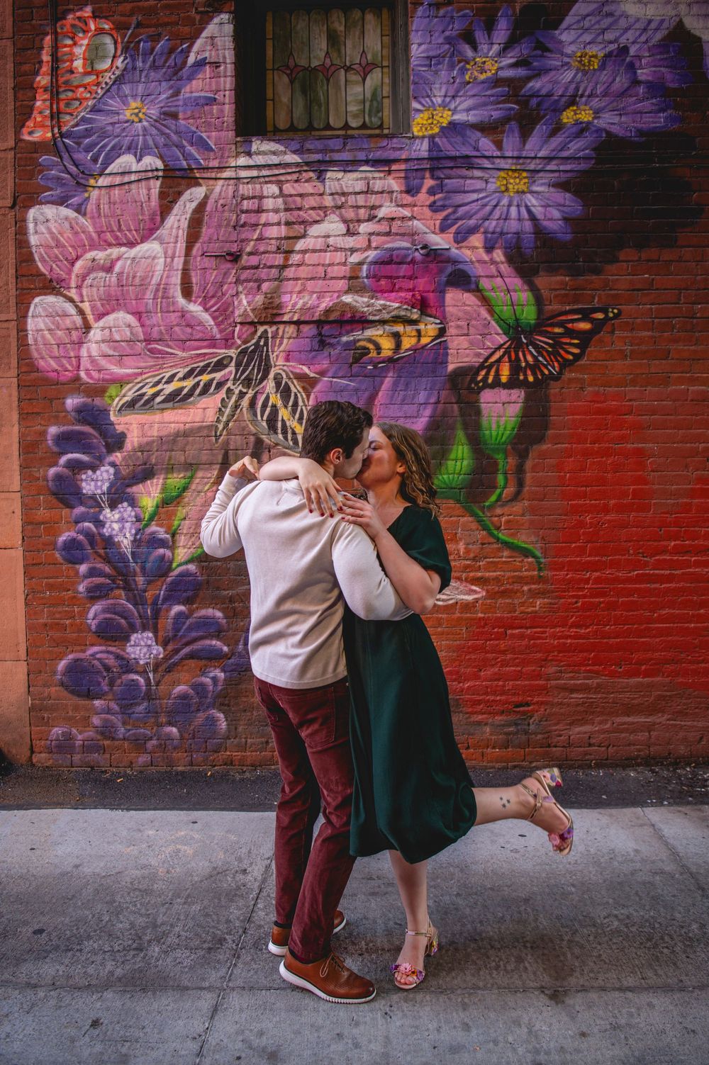 Couple sharing a romantic kiss in front of a colorful floral butterfly mural on a brick wall.