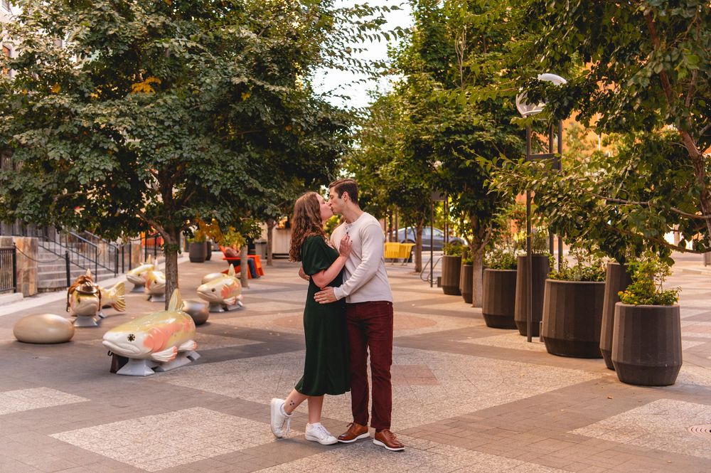 Couple sharing a romantic moment on a tree-lined pedestrian plaza with decorative fish sculptures.