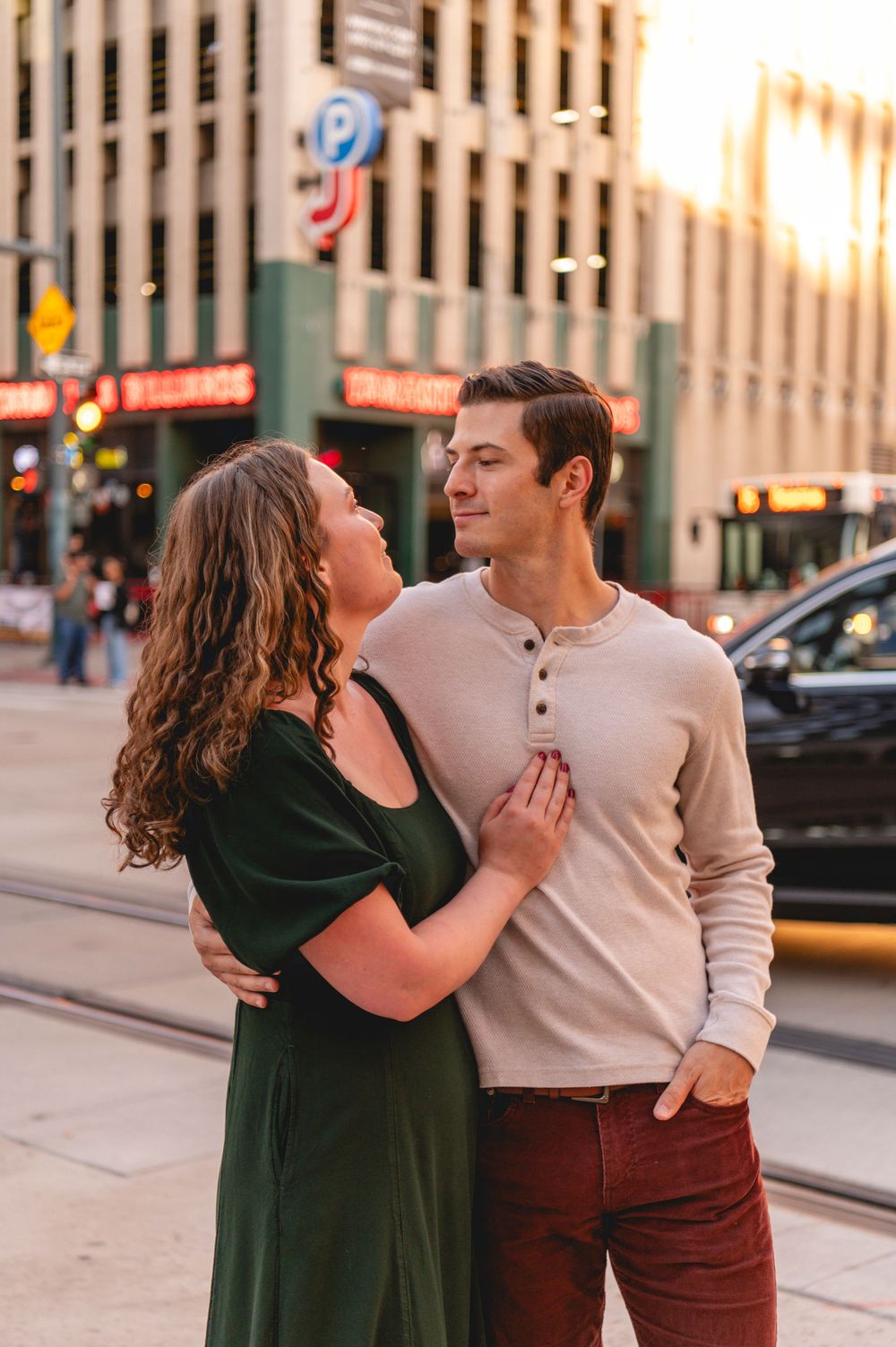 A couple embraces on a city street at golden hour, the woman in green dress gazing up at the man in a beige sweater.