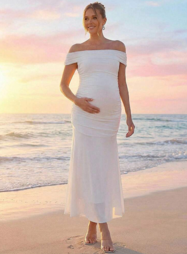 Pregnant woman in white off-shoulder ruched maxi dress posing on beach at sunset.