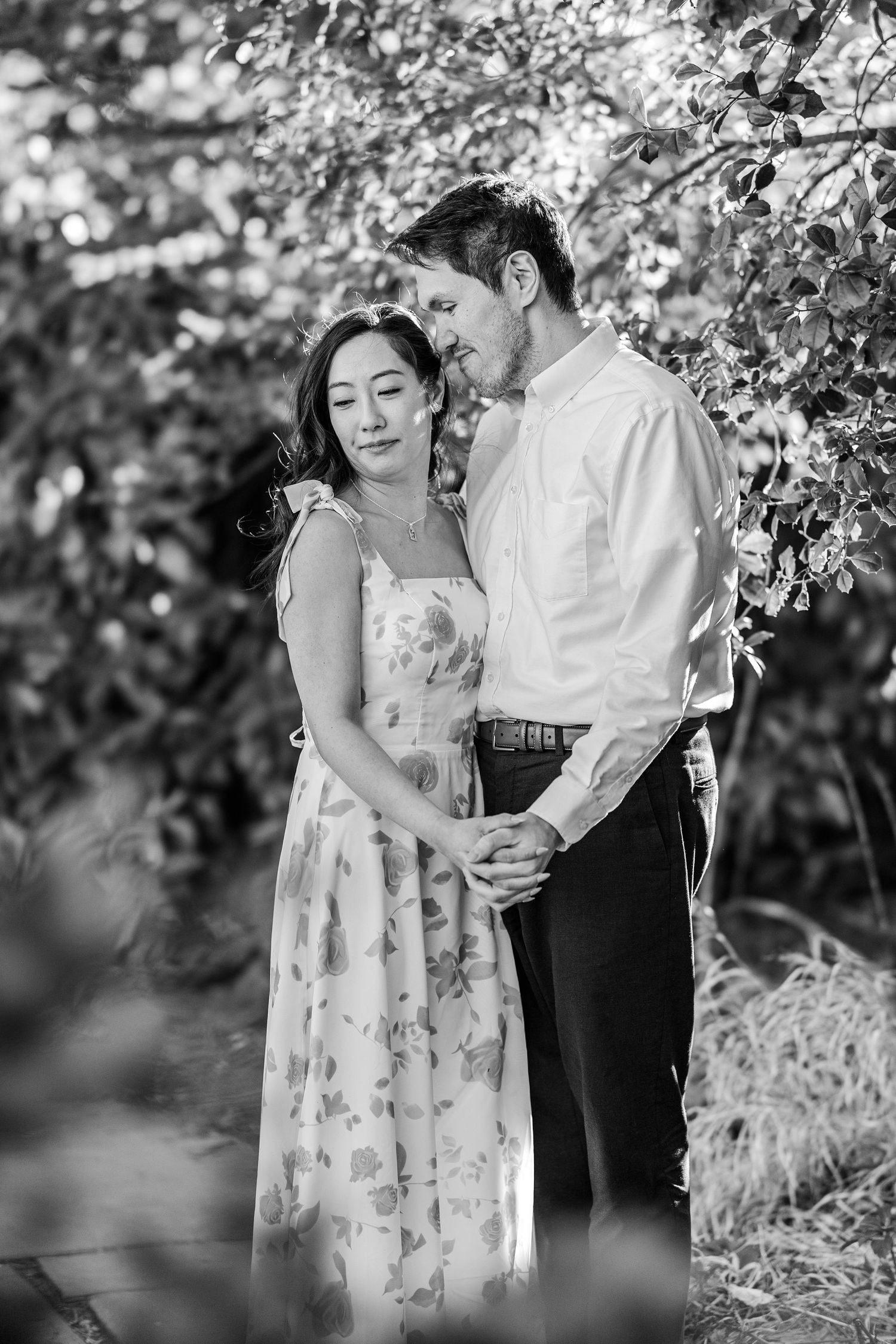 Black and white photo of a couple embracing tenderly in a lush garden, woman in floral dress, man in white shirt.