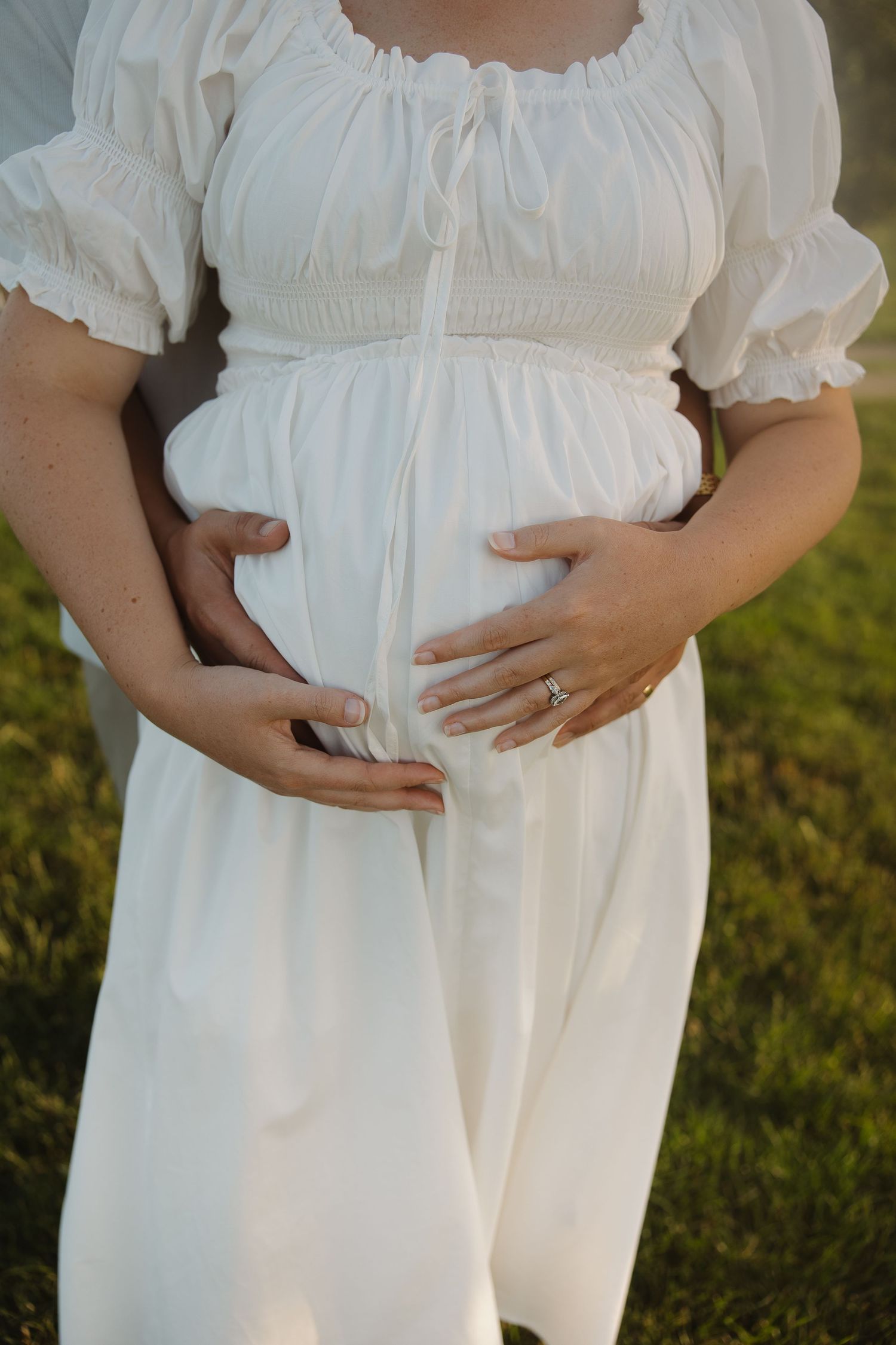 Pregnant woman in white smocked dress cradling baby bump outdoors, wearing engagement ring, standing on green grass.