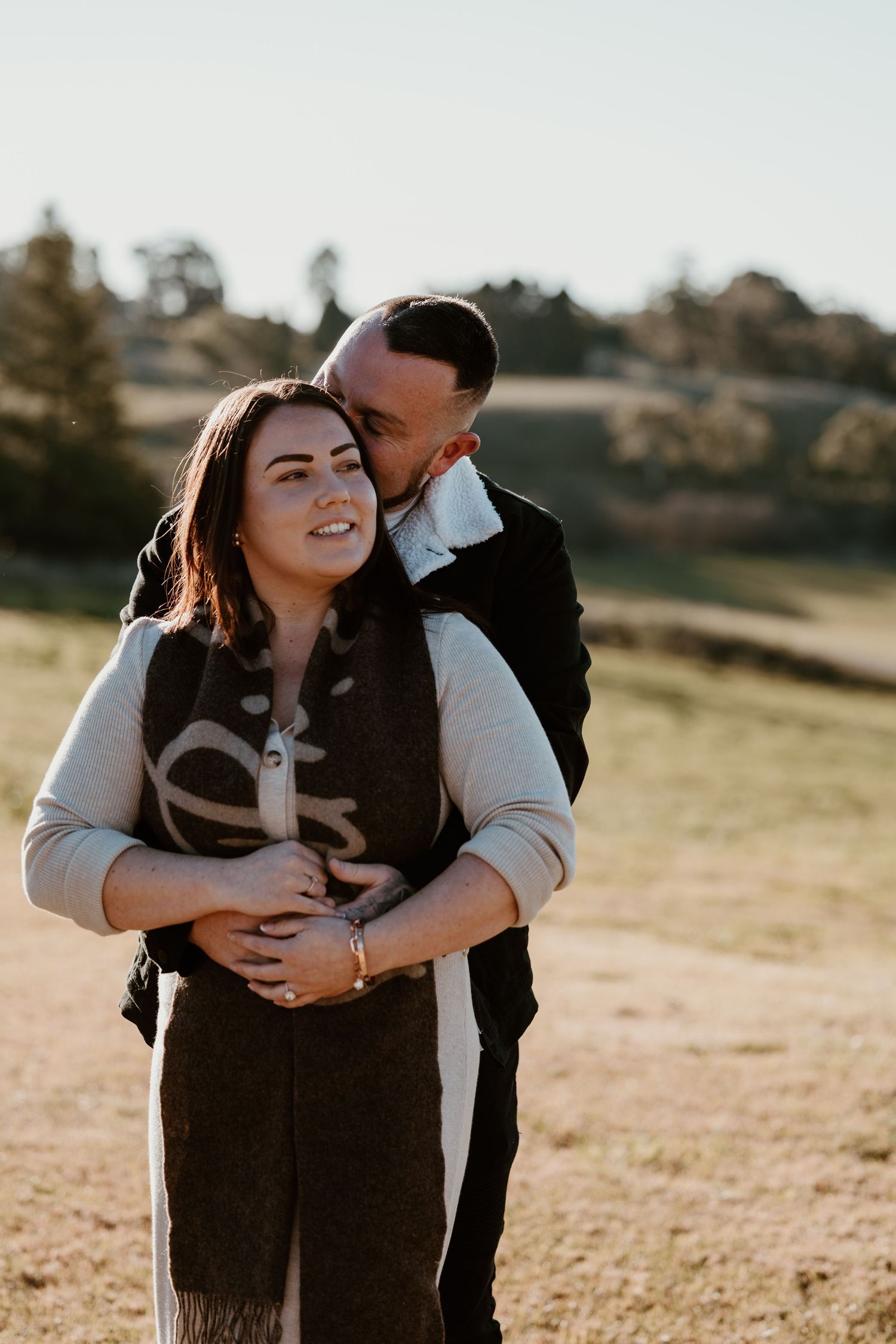 Couple embracing outdoors at golden hour, man kissing woman's cheek in a sunlit field with rolling hills behind them.