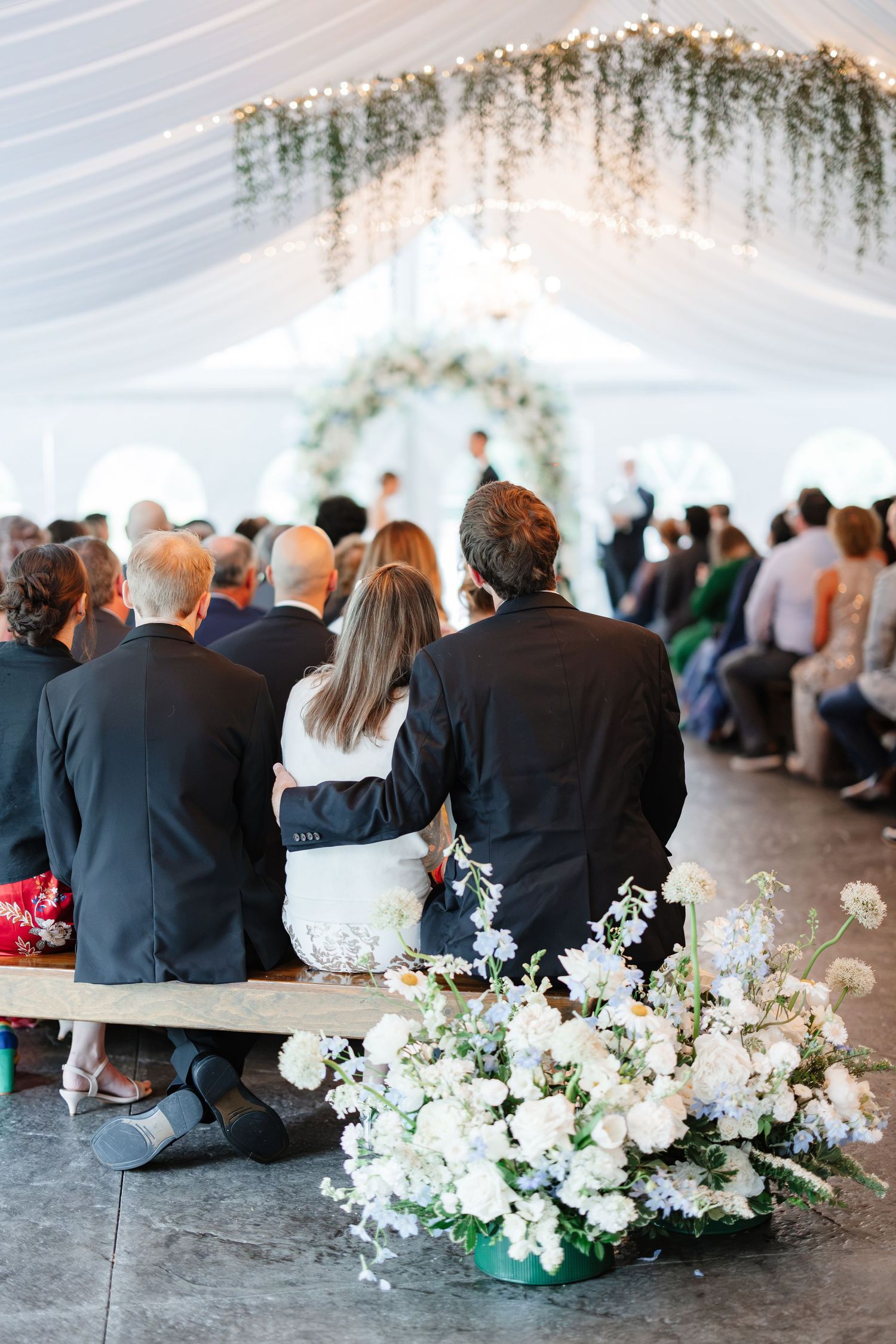 Wedding guests seated on benches watching ceremony beneath draped tent with white floral arch.