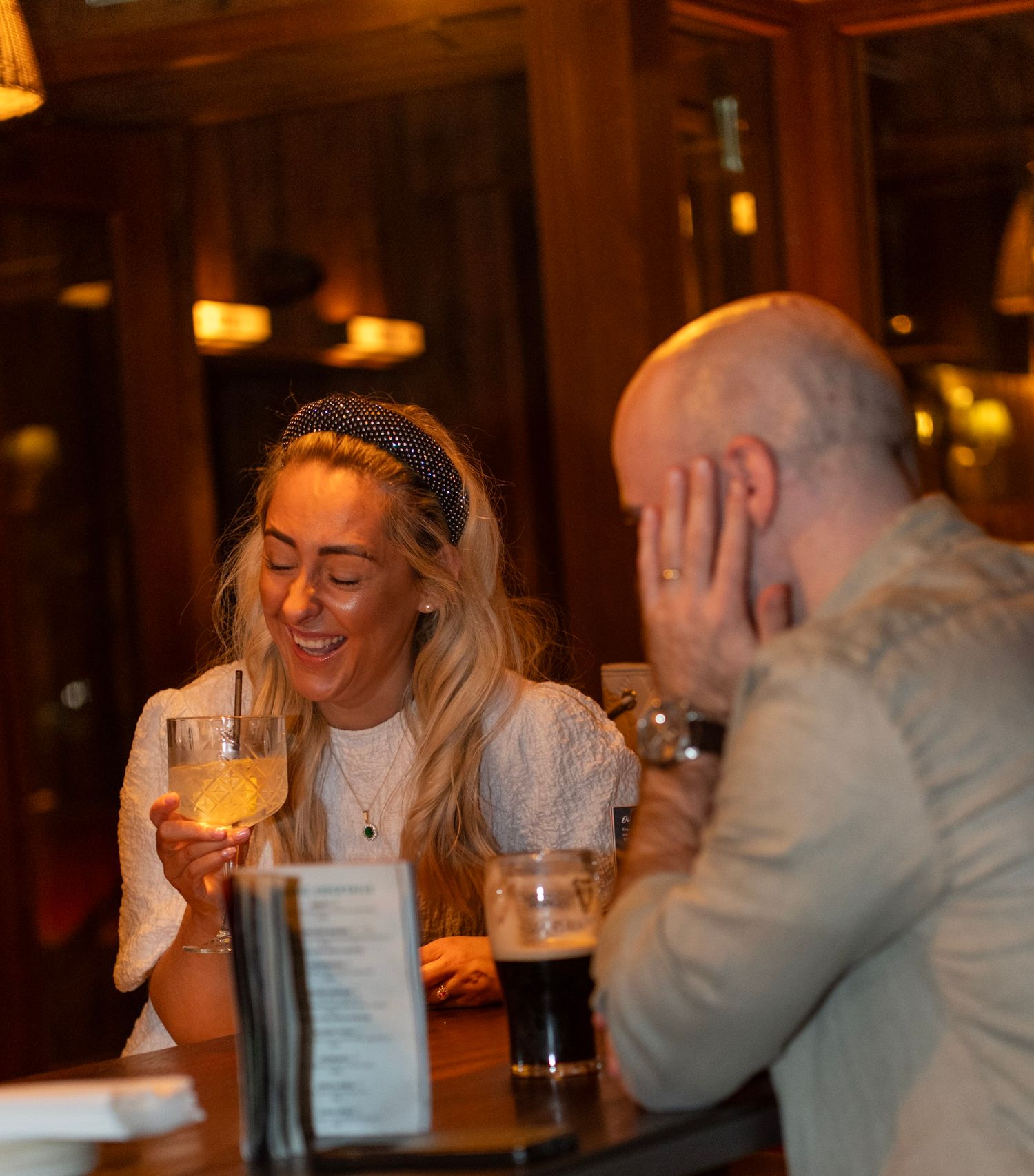 Couple laughing and enjoying drinks at a cozy bar, woman holding white wine glass, warm ambient lighting.