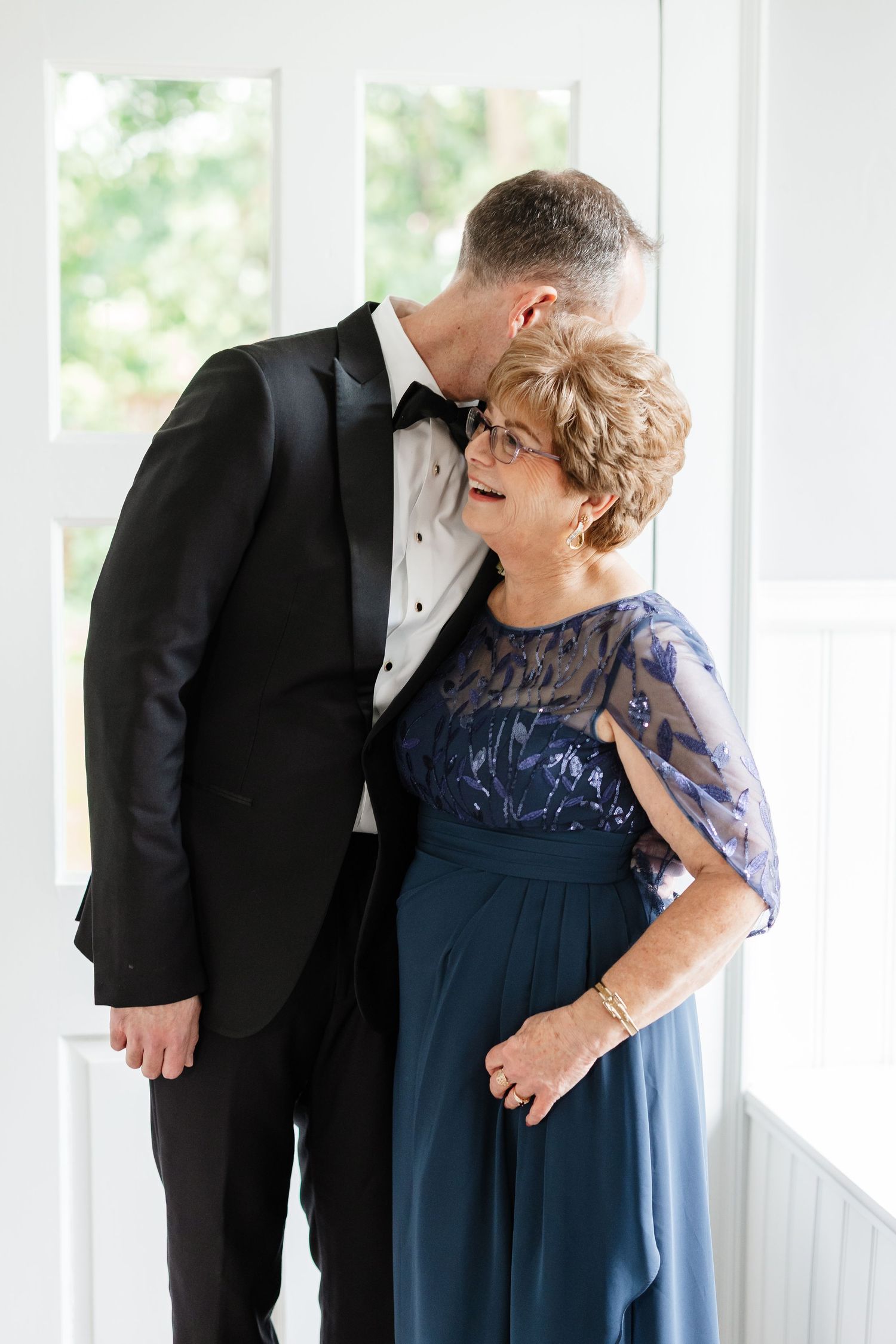 Groom in black tuxedo sharing tender moment with mother in navy blue lace gown at wedding venue.