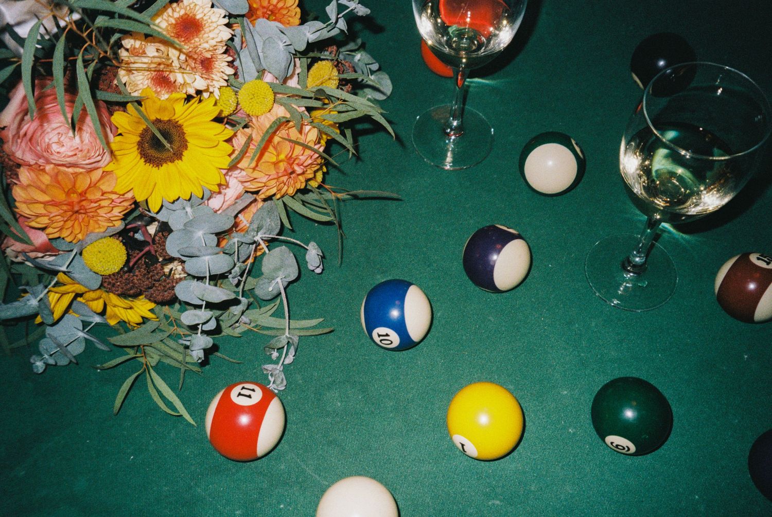 Colorful billiard balls and wine glasses arranged on a green pool table beside a vibrant floral bouquet.