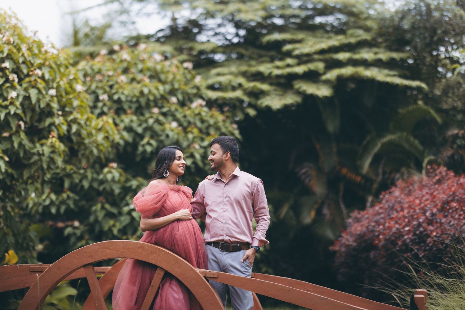 Couple poses on a wooden bridge surrounded by lush bamboo trees, woman holding baby shoes during maternity photoshoot.